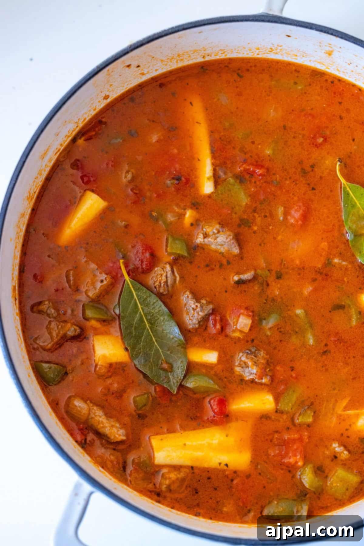 A pot of beef stew overflowing with tender chunks of meat, yellow potatoes, tomatoes, green bell peppers, and bay leaves, all immersed in a rich, spiced broth.