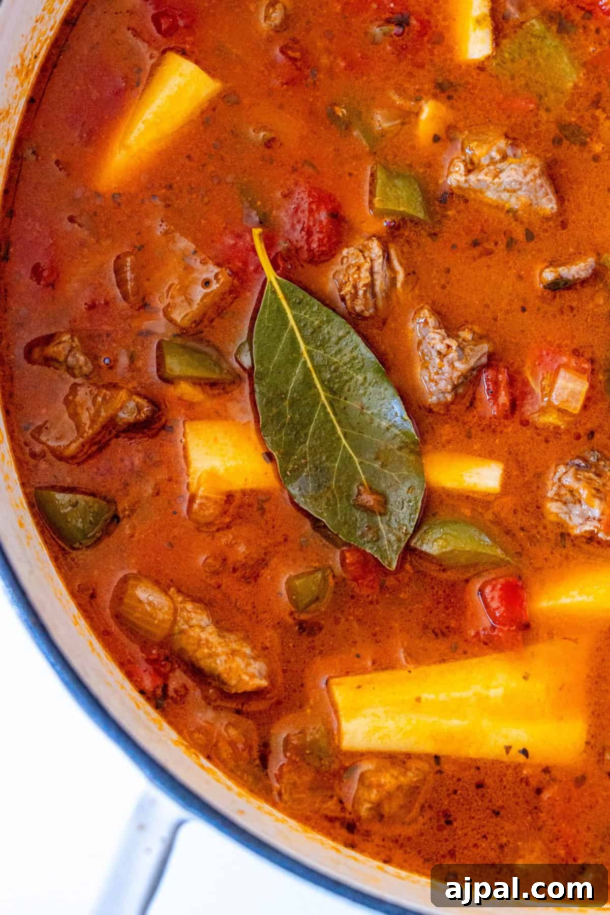 A large pot of hearty beef stew, featuring tender chunks of beef, potatoes, parsnip, green peppers, tomatoes, and a bay leaf, simmering in a rich, red broth, ready to be served.