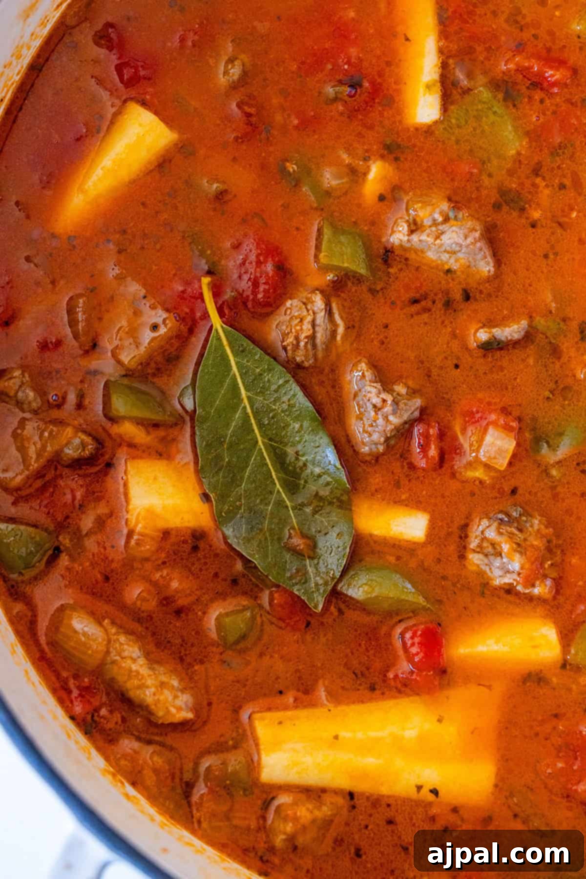 A close-up view of a simmering beef and parsnip stew, showcasing tender chunks of beef, vibrant green bell peppers, diced tomatoes, potatoes, parsnip slices, and an aromatic bay leaf, all blending in a rich broth.
