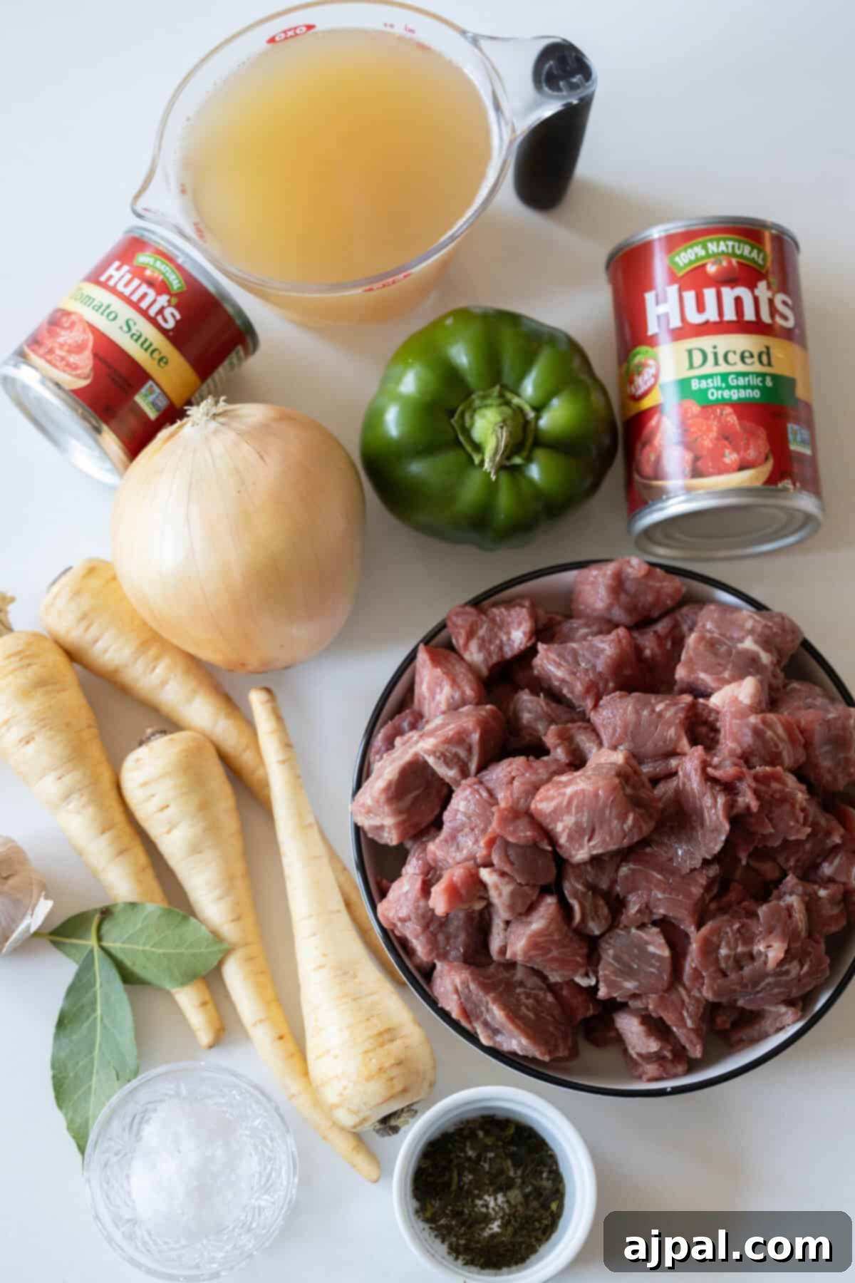 A vibrant assortment of stew ingredients laid out: raw beef cubes, aromatic onion, crisp green bell pepper, and sweet parsnips. Canned tomatoes, rich beef broth, and fragrant bay leaves are also present, arranged on a light surface for a harmonious blend of flavors.
