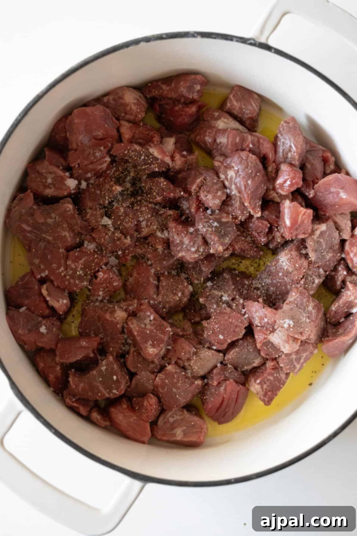 Chunks of raw beef and parsnip, seasoned with salt and pepper, begin to sizzle in a white pot with olive oil, ready for browning.