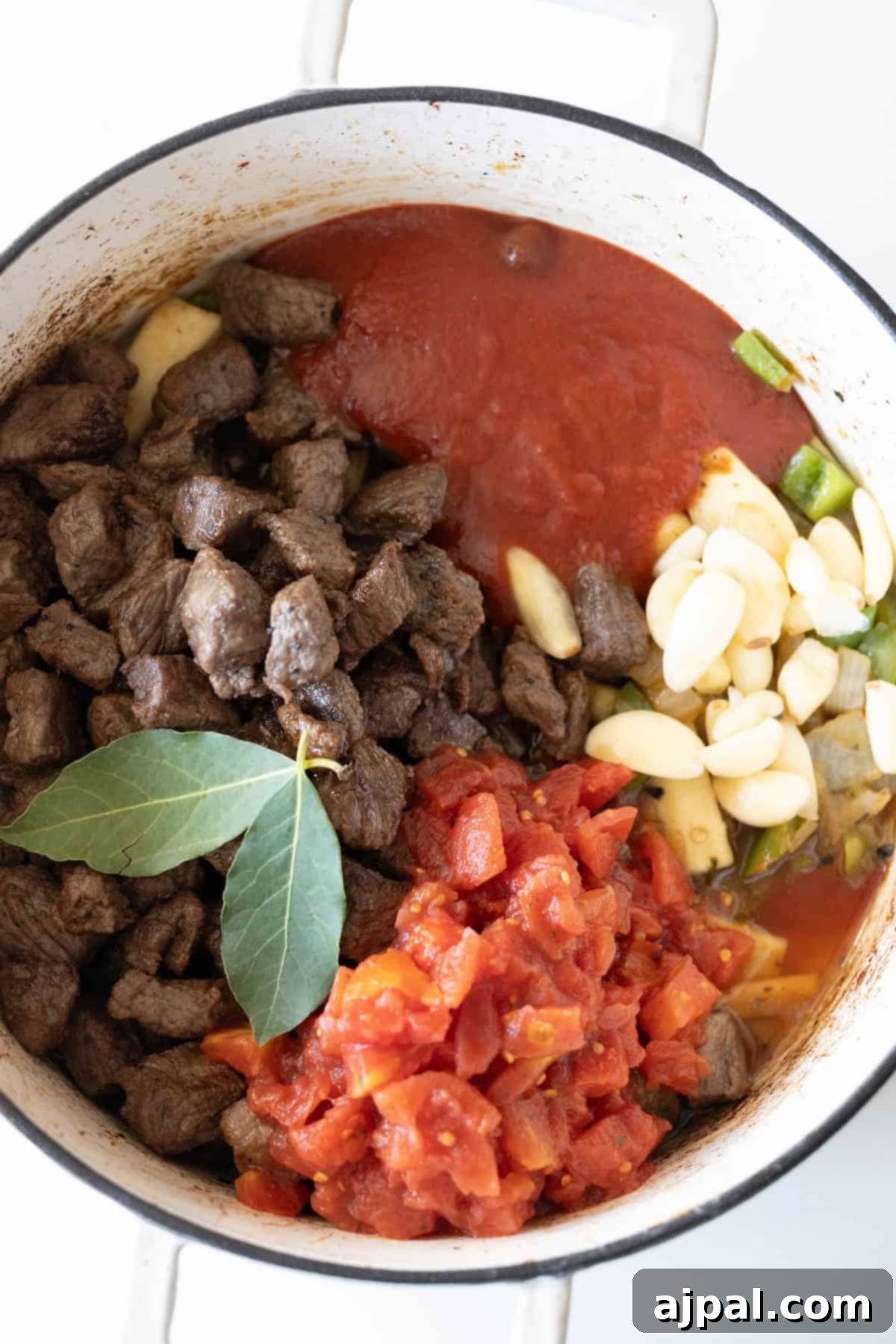 A pot of beef stew with tender cooked beef, green peppers, sliced garlic, and diced tomatoes in a rich tomato sauce, enhanced by a fragrant bay leaf.