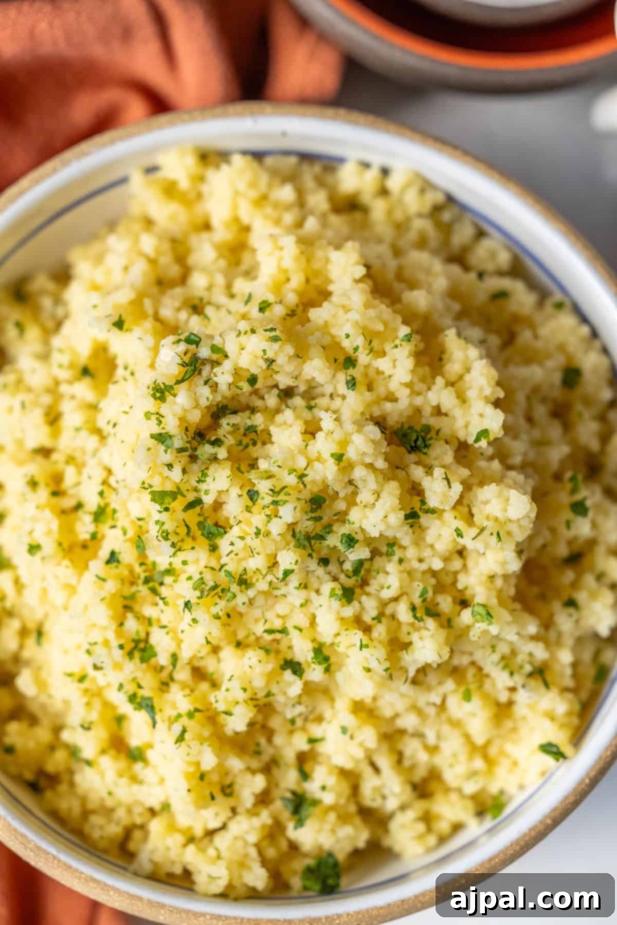 Authentic Couscous Delights 2 A bowl filled with perfectly cooked couscous is garnished with fresh chopped herbs, offering a delightful recipe experience. An orange cloth is partially visible next to the bowl.