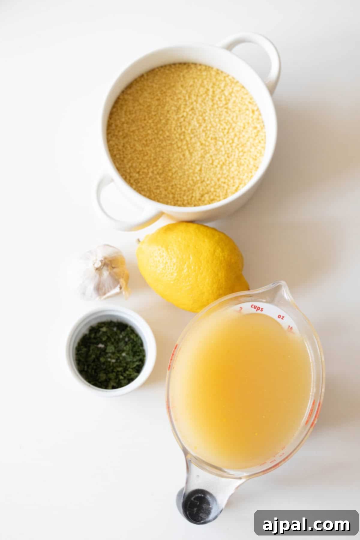 Authentic Couscous Delights 5 A bowl of Cous Cous, a lemon, a garlic bulb, chopped herbs, and a measuring cup of broth on a white surface set the stage for your next culinary adventure.