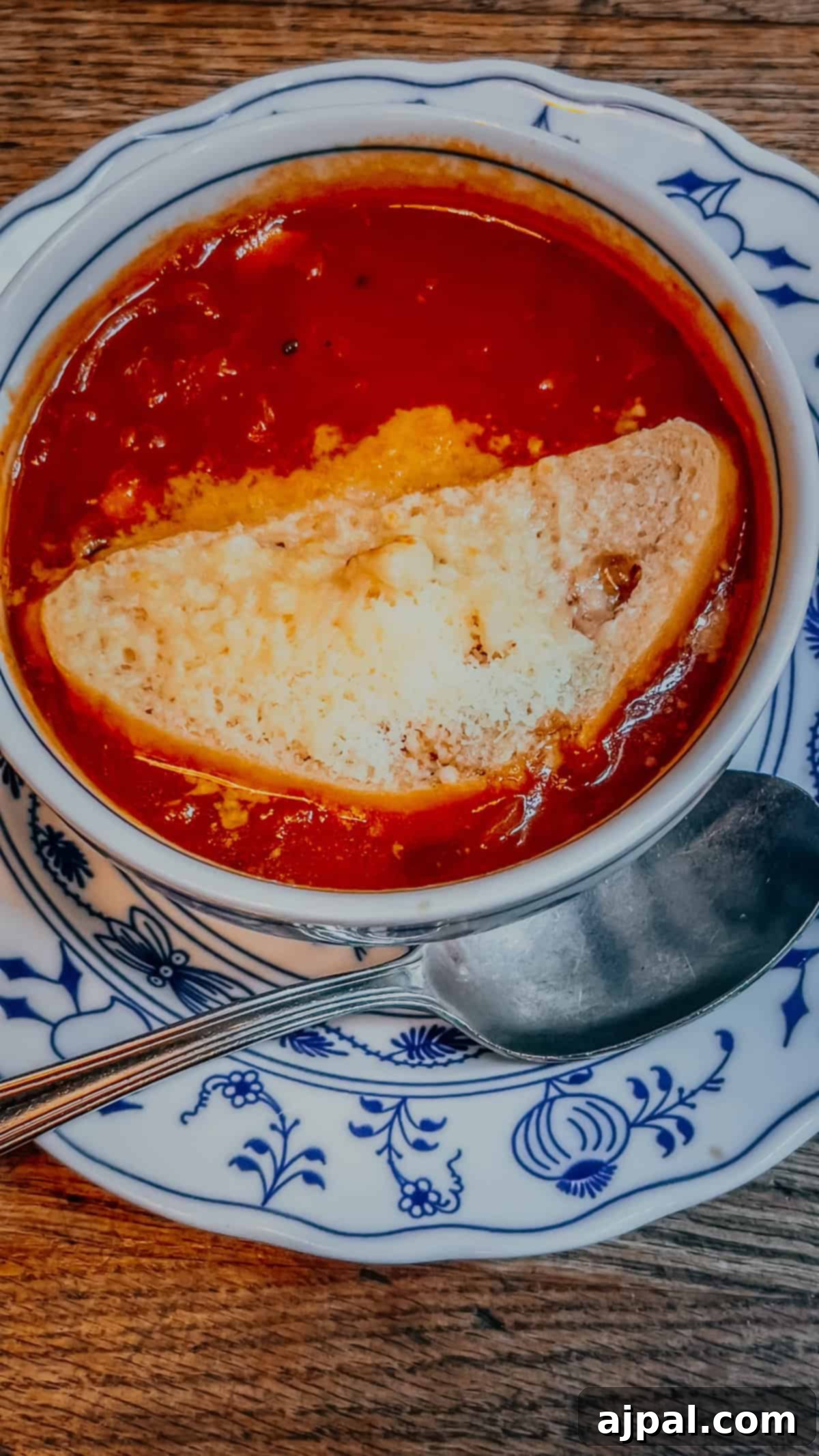 Bolzano's Magical Christmas Market 11 A bowl of tomato soup with a slice of bread and melted cheese on top, reminiscent of the cozy flavors found at a Christmas Market in Bolzano, served on a floral-patterned plate with a spoon beside it.