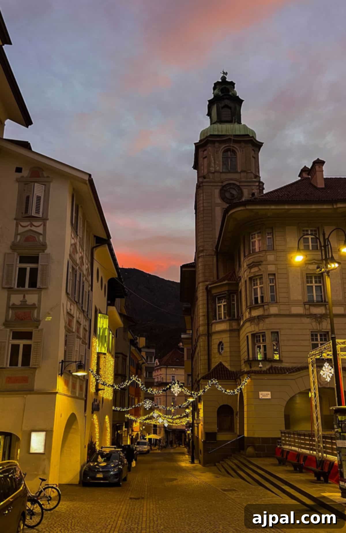 Bolzano's Magical Christmas Market 16 A narrow cobblestone street lined with buildings and festive lights, reminiscent of a cozy Christmas market in Bolzano, features a clock tower set against a colorful sunset sky.
