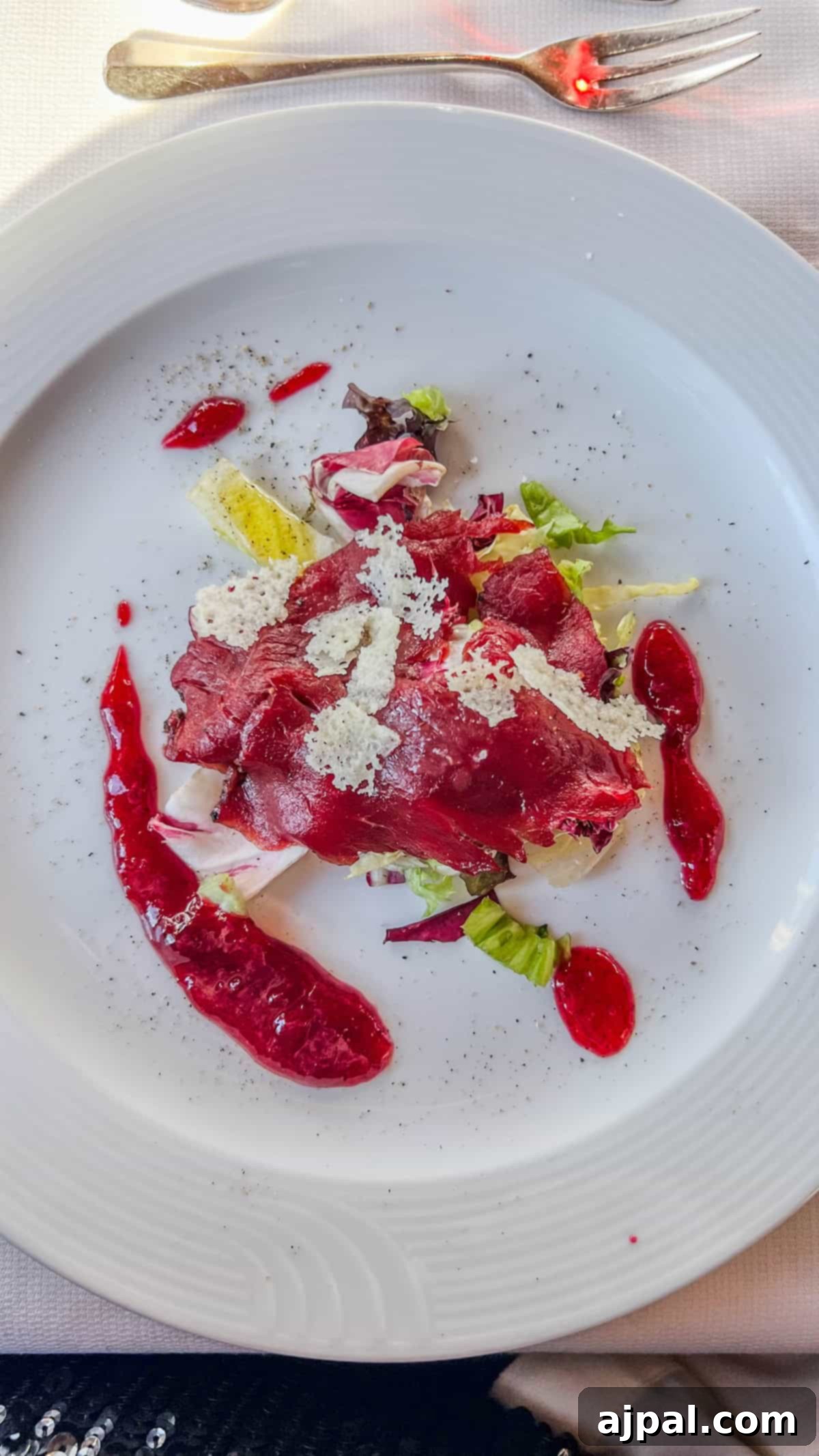Bolzano's Magical Christmas Market 18 A plate of sliced beetroot with green lettuce, white cheese shavings, and red sauce drizzled on the side evokes the festive spirit of a Christmas Market in Bolzano. The vibrant dish is elegantly presented on a white plate, with a fork placed at the top right.