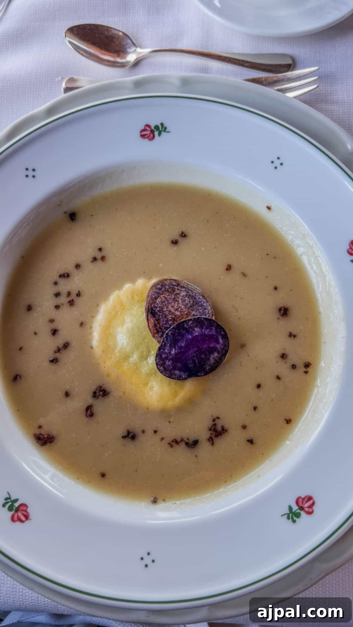 Bolzano's Magical Christmas Market 19 A bowl of creamy soup topped with two purple potato slices and a yellow dollop sits on a decorative white dish with a floral pattern, reminiscent of the elegance found at Bolzano's Christmas Market. Silverware is placed beside the plate.
