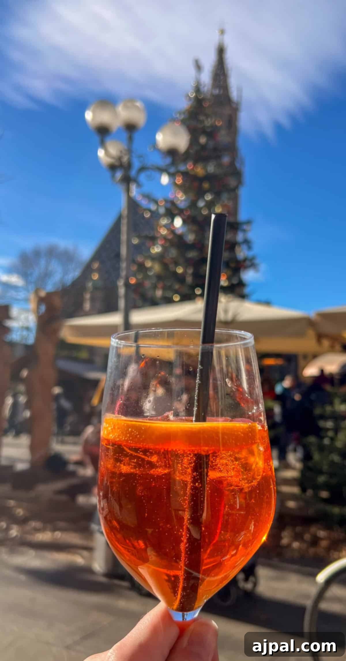 Bolzano's Magical Christmas Market 4 A glass of orange cocktail with a straw is held up in an outdoor setting, with a blurred view of a Christmas tree and the charming buildings of Bolzano's market in the background.