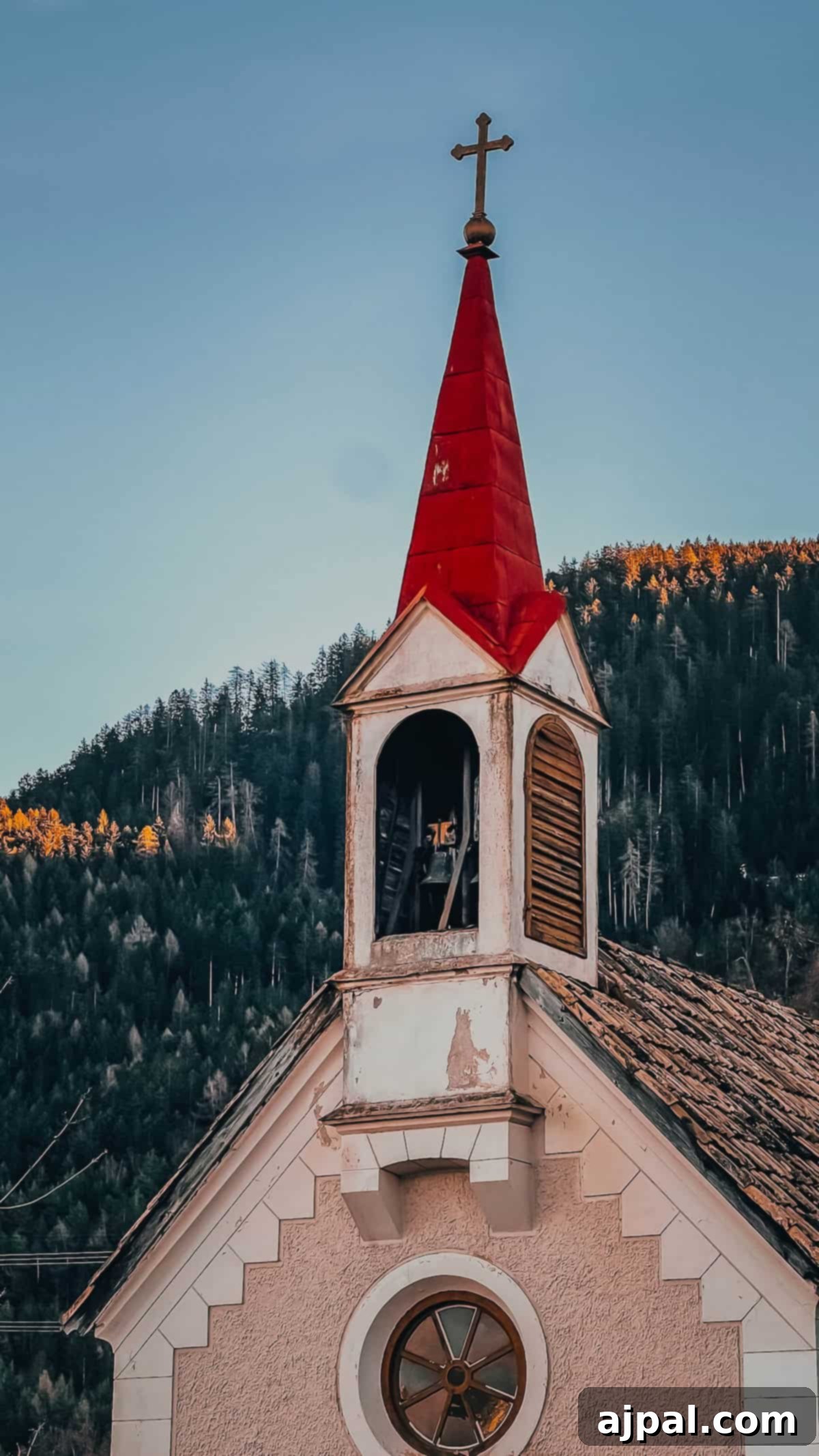 Bolzano's Magical Christmas Market 7 Nestled near Bolzano, this small church with a red steeple and cross stands serenely against forested hills under a clear sky, evoking the charm of a Christmas market scene.