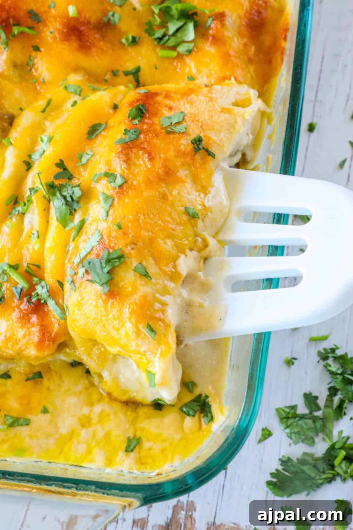 Ultimate Creamy White Chicken Enchiladas 11 A baked cheesy casserole is being served with a white spatula. The dish is garnished with fresh parsley and features a golden-brown top layer.