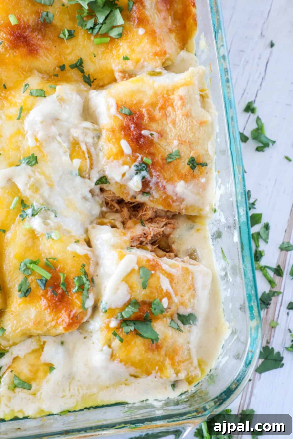 Ultimate Creamy White Chicken Enchiladas 13 Close-up of baking dish with enchiladas topped with melted cheese and chopped cilantro, featuring a white cream sauce and shredded chicken filling, on a wooden table.