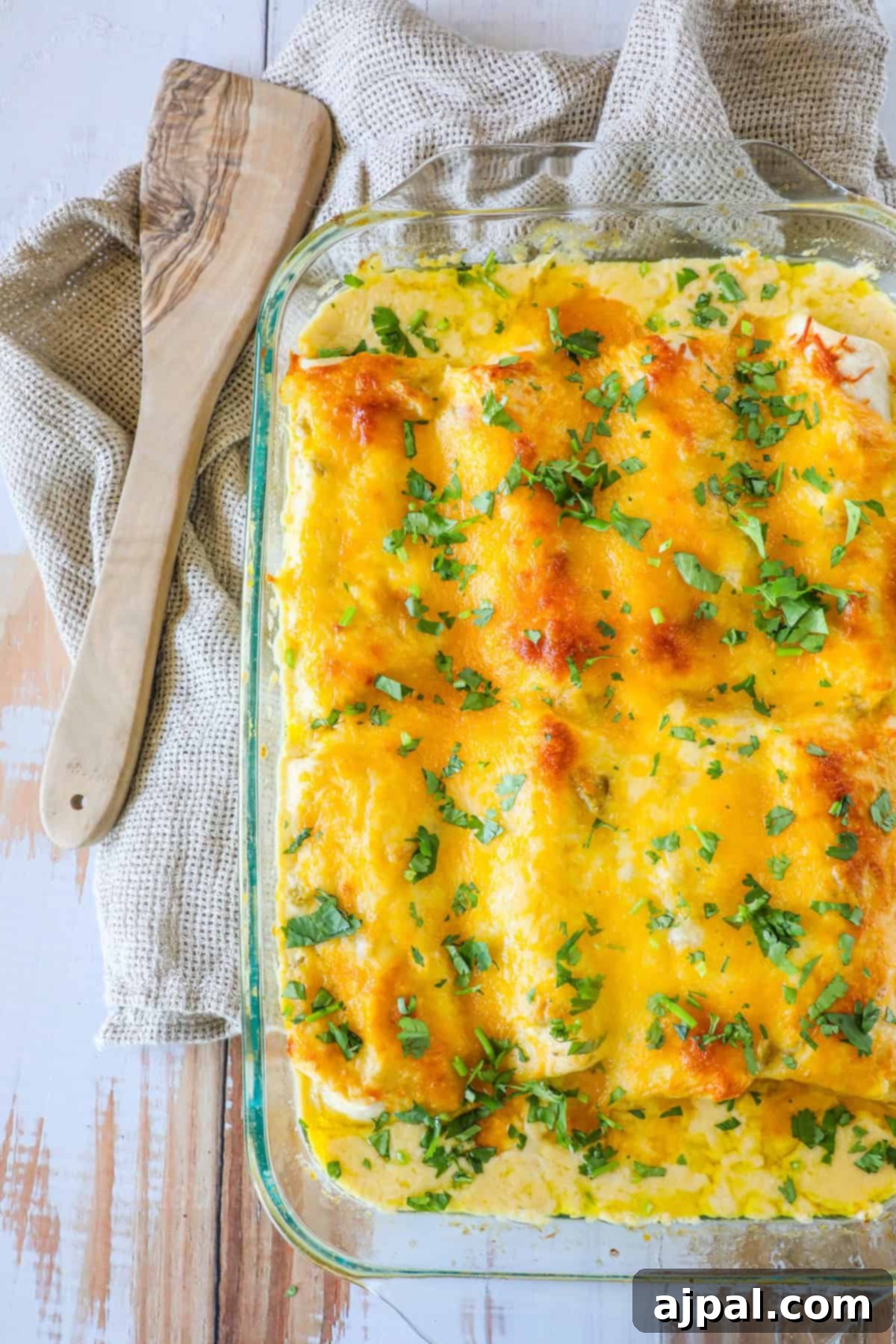 Ultimate Creamy White Chicken Enchiladas 14 A glass baking dish filled with baked, cheesy enchiladas topped with chopped green herbs. A wooden spatula rests on a beige cloth beside the dish.