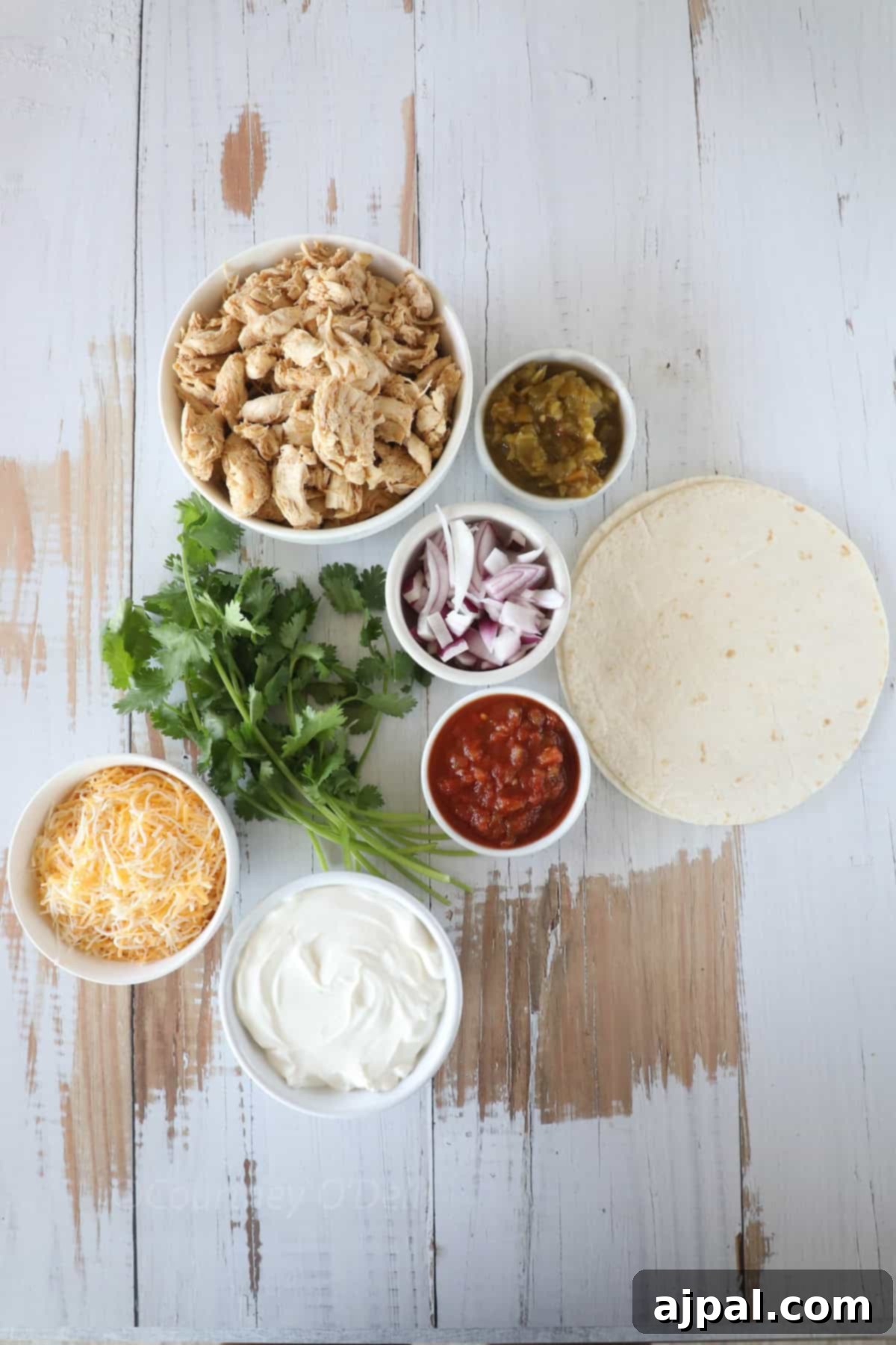 Ultimate Creamy White Chicken Enchiladas 3 Ingredients for a meal: shredded chicken, chopped cilantro, shredded cheese, sour cream, diced onions, salsa, green chilies, and flour tortillas arranged on a wooden surface.