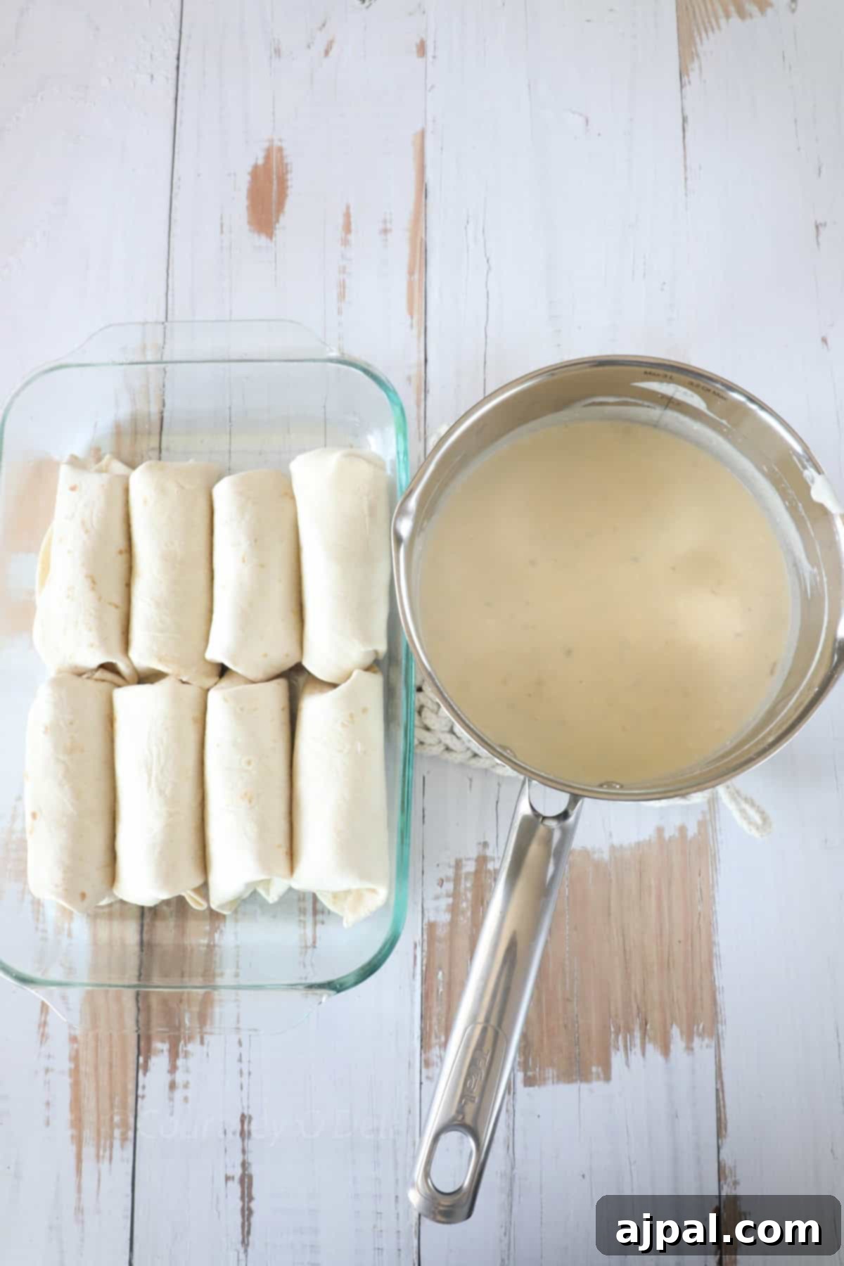 Ultimate Creamy White Chicken Enchiladas 4 A glass baking dish with eight rolled tortillas sits next to a saucepan containing a light-colored sauce on a weathered wooden surface.