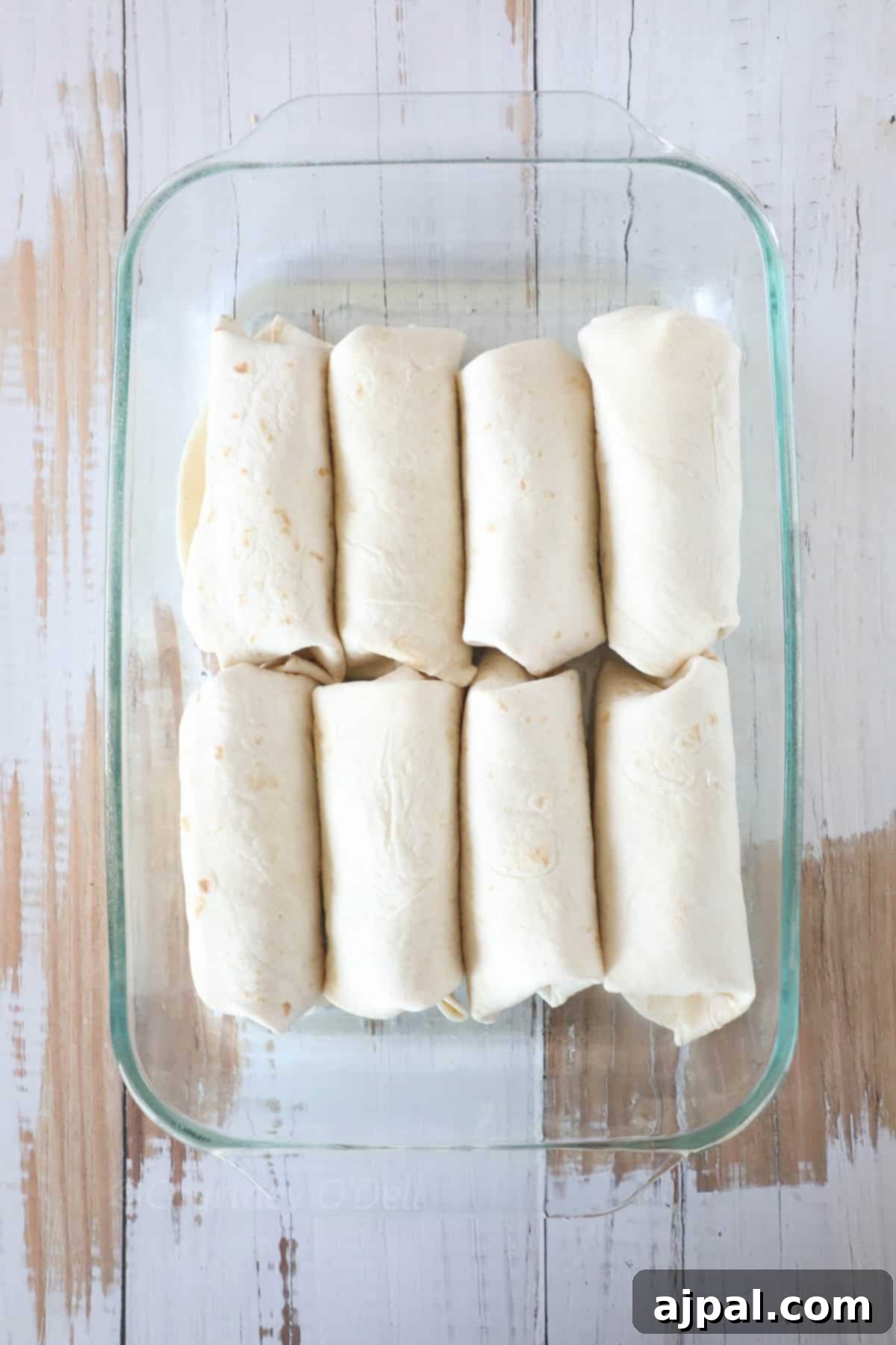 Ultimate Creamy White Chicken Enchiladas 7 Eight uncooked burritos are arranged in a glass baking dish on a wooden surface.