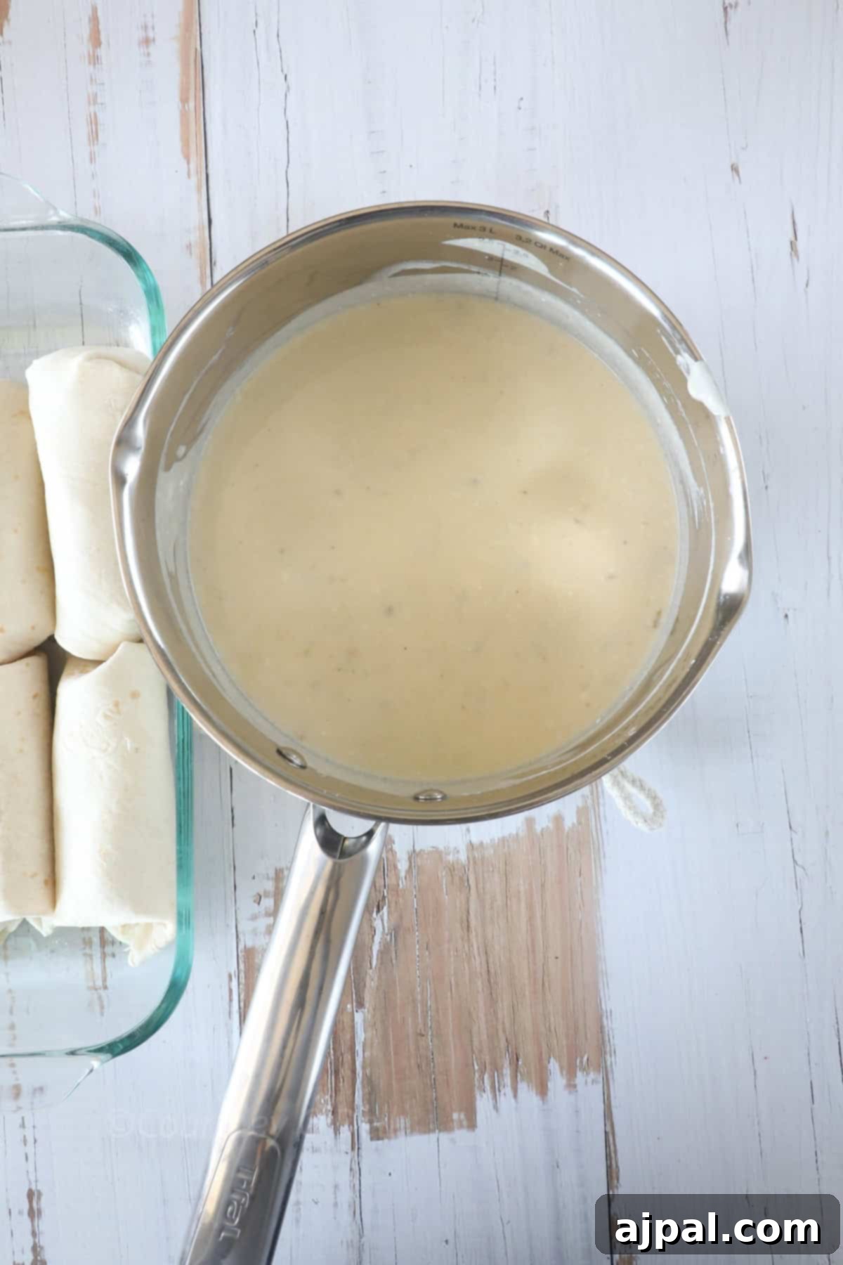 Ultimate Creamy White Chicken Enchiladas 8 A saucepan filled with creamy sauce is placed on a white wooden surface. Next to it, a glass dish contains several rolled tortillas.