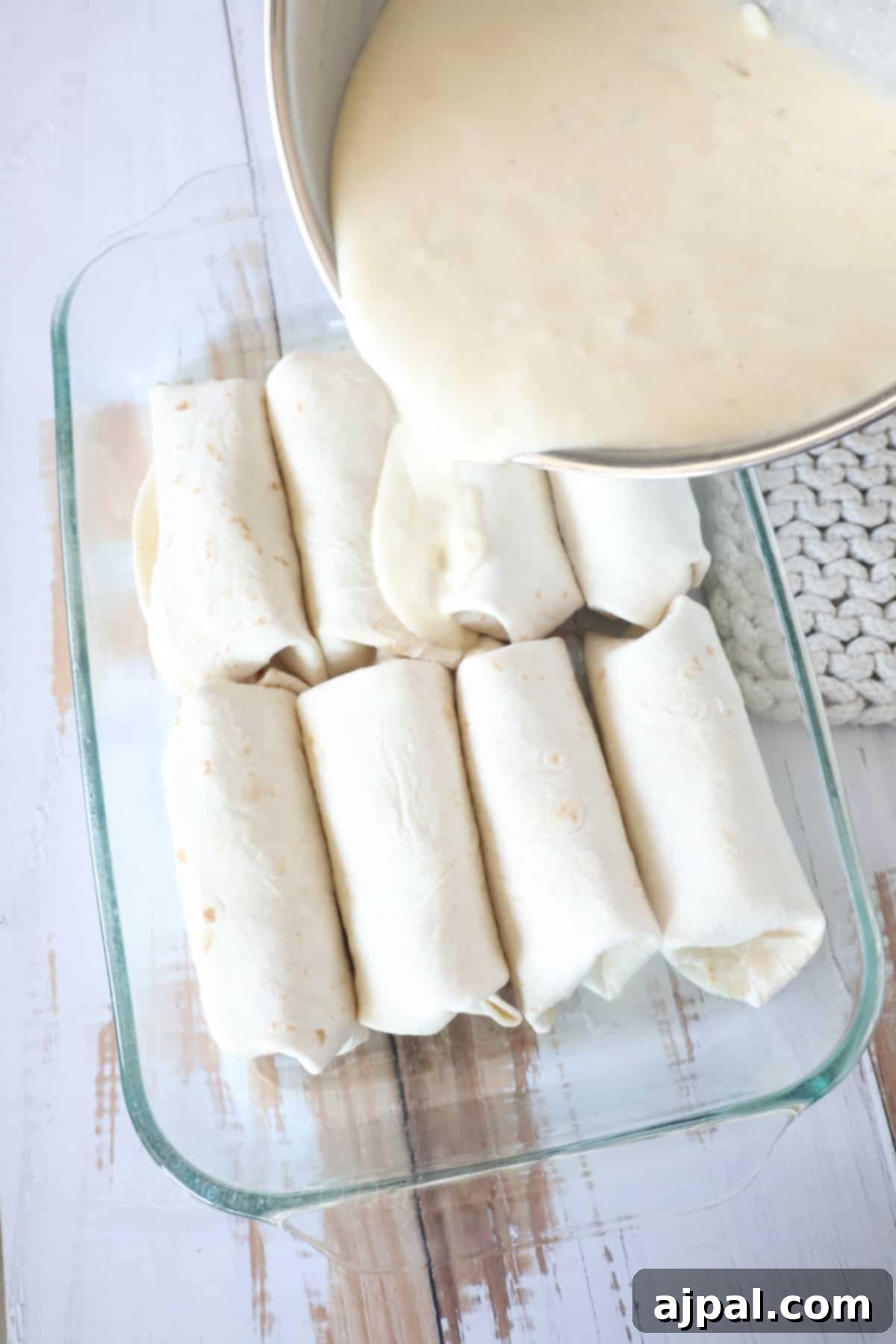 Ultimate Creamy White Chicken Enchiladas 9 A glass baking dish with rolled tortillas is being covered in a white sauce poured from a pot.