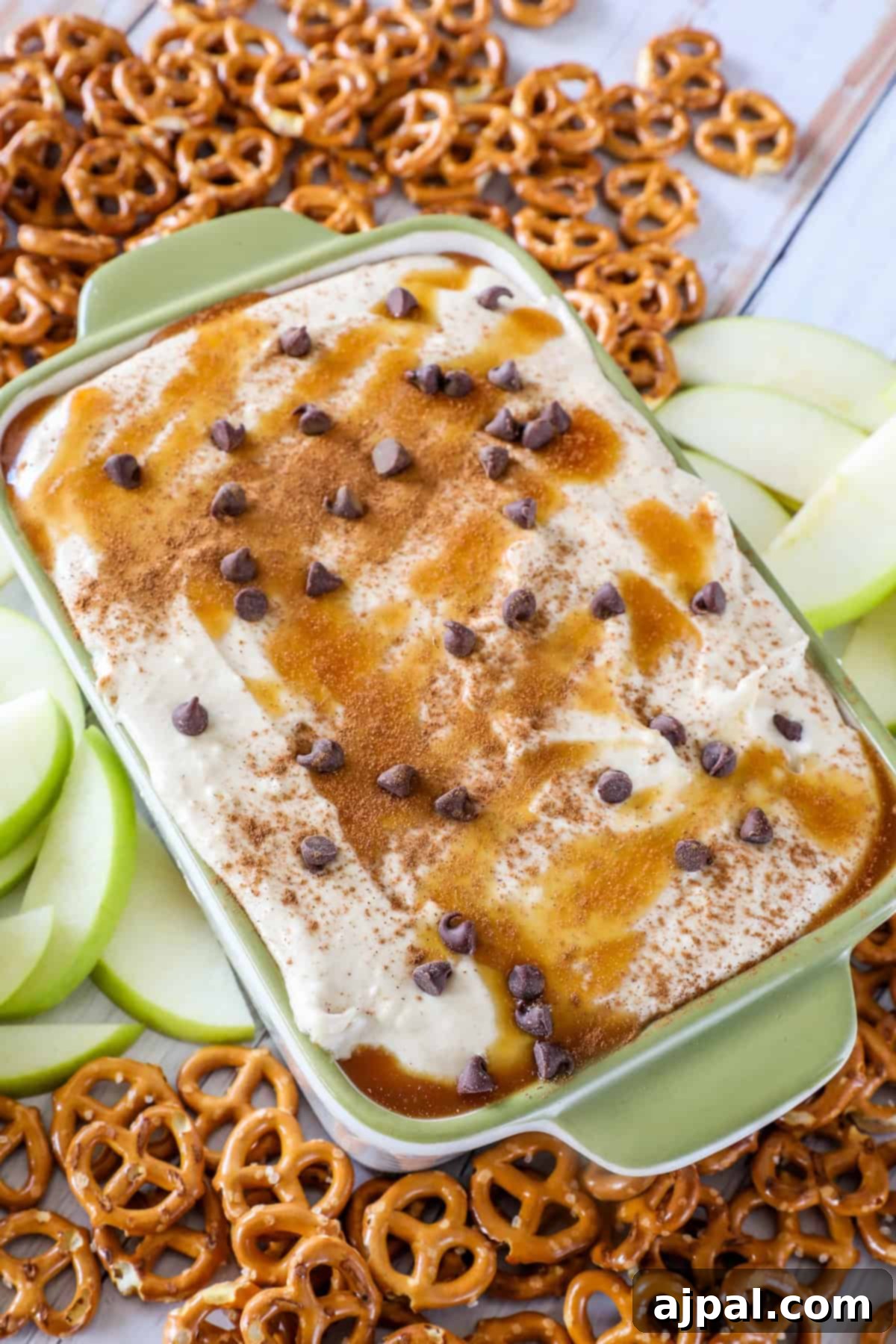 A green dish with creamy caramel apple dip topped with chocolate chips and cinnamon, surrounded by apple slices and pretzels.