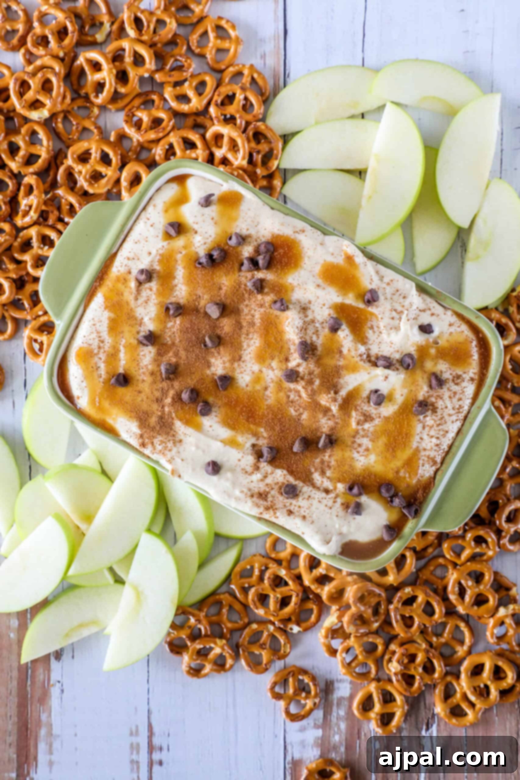 Caramel apple dip topped with chocolate chips in a dish, surrounded by sliced apples and pretzels on a wooden surface.