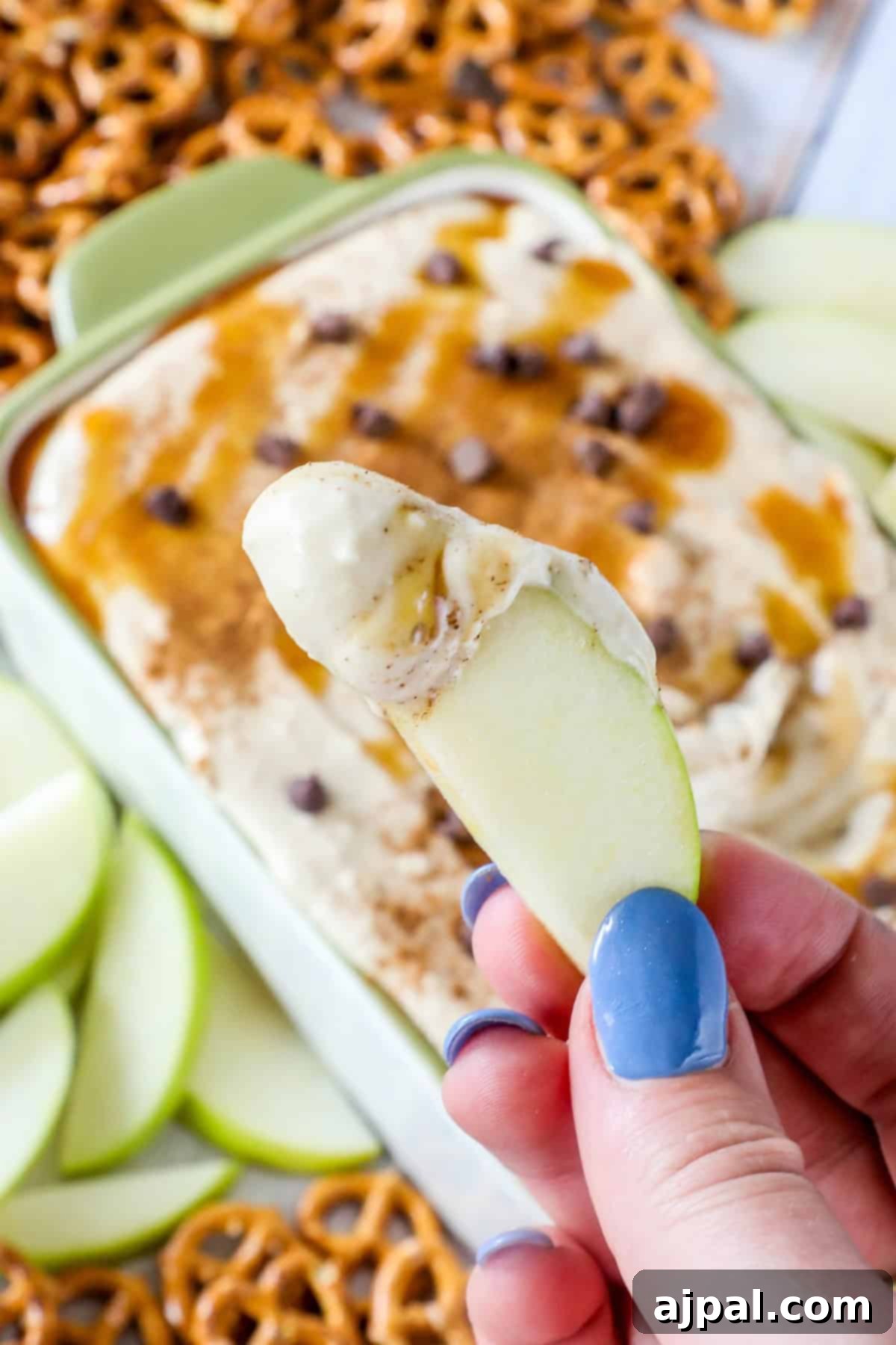 A person holding a slice of green apple dipped in a rich caramel apple dip, with pretzels and more apple slices visible in the background.