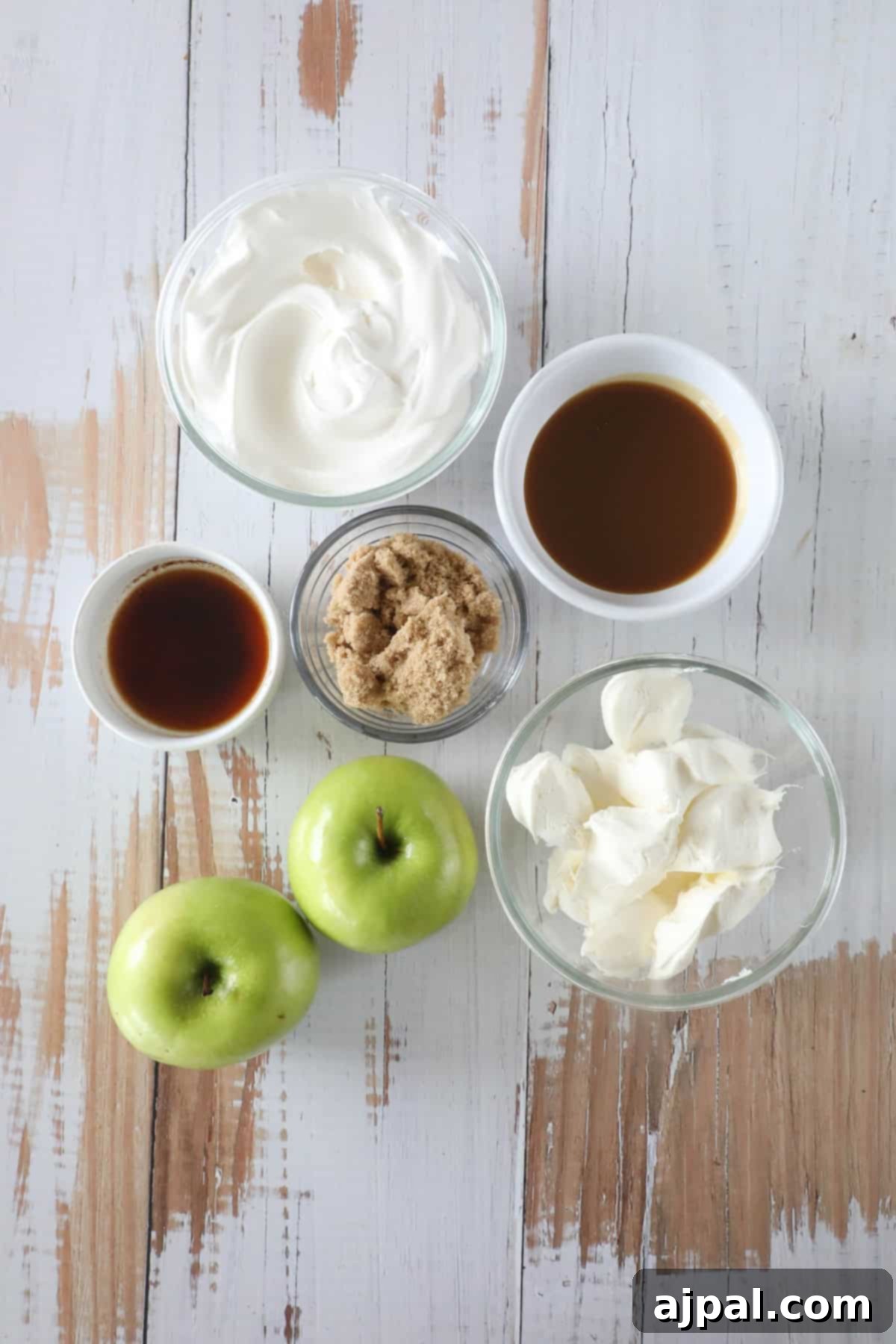 On a wooden surface, ingredients for a caramel apple dip include two green apples, bowls of brown sugar, cream cheese, rich caramel, vanilla extract, and fluffy whipped cream.