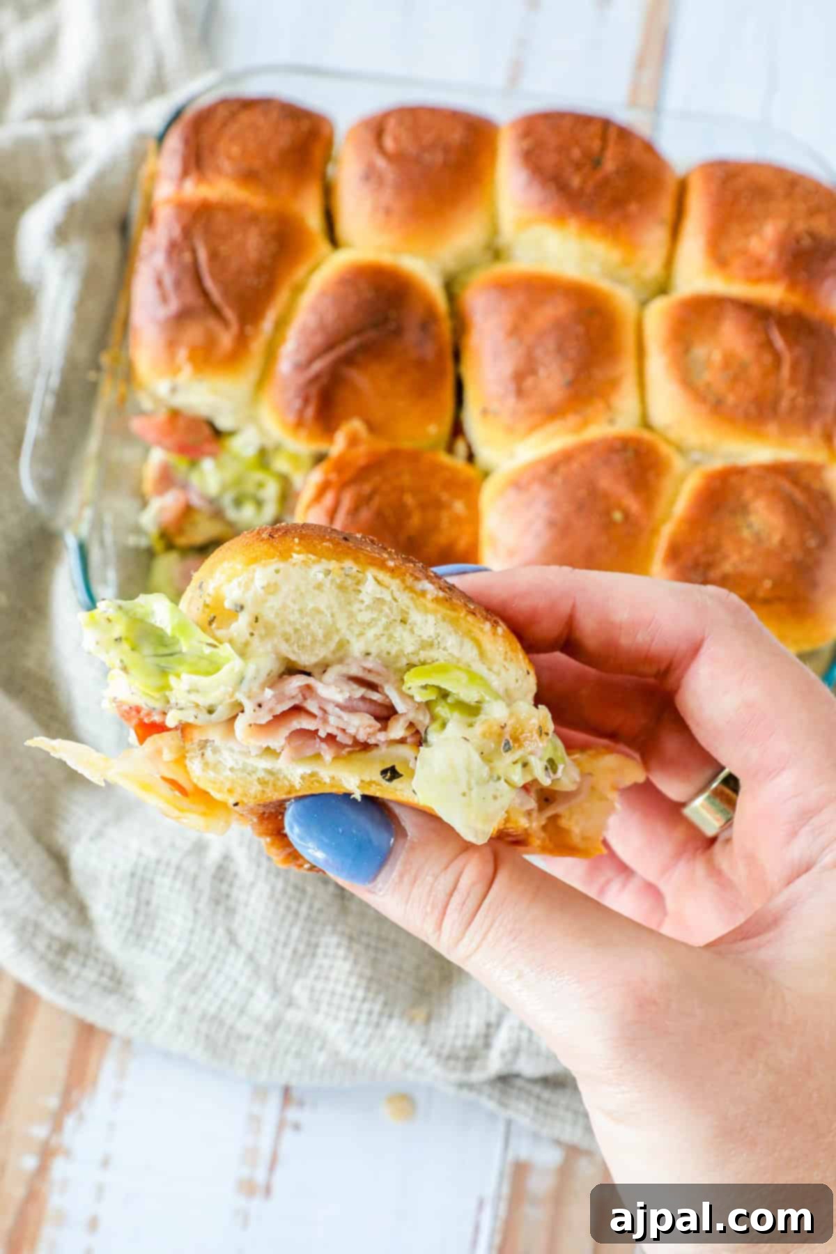 A hand holding a slider with lettuce, bacon, and toppings. A tray of similar sliders is in the background on a wooden surface.
