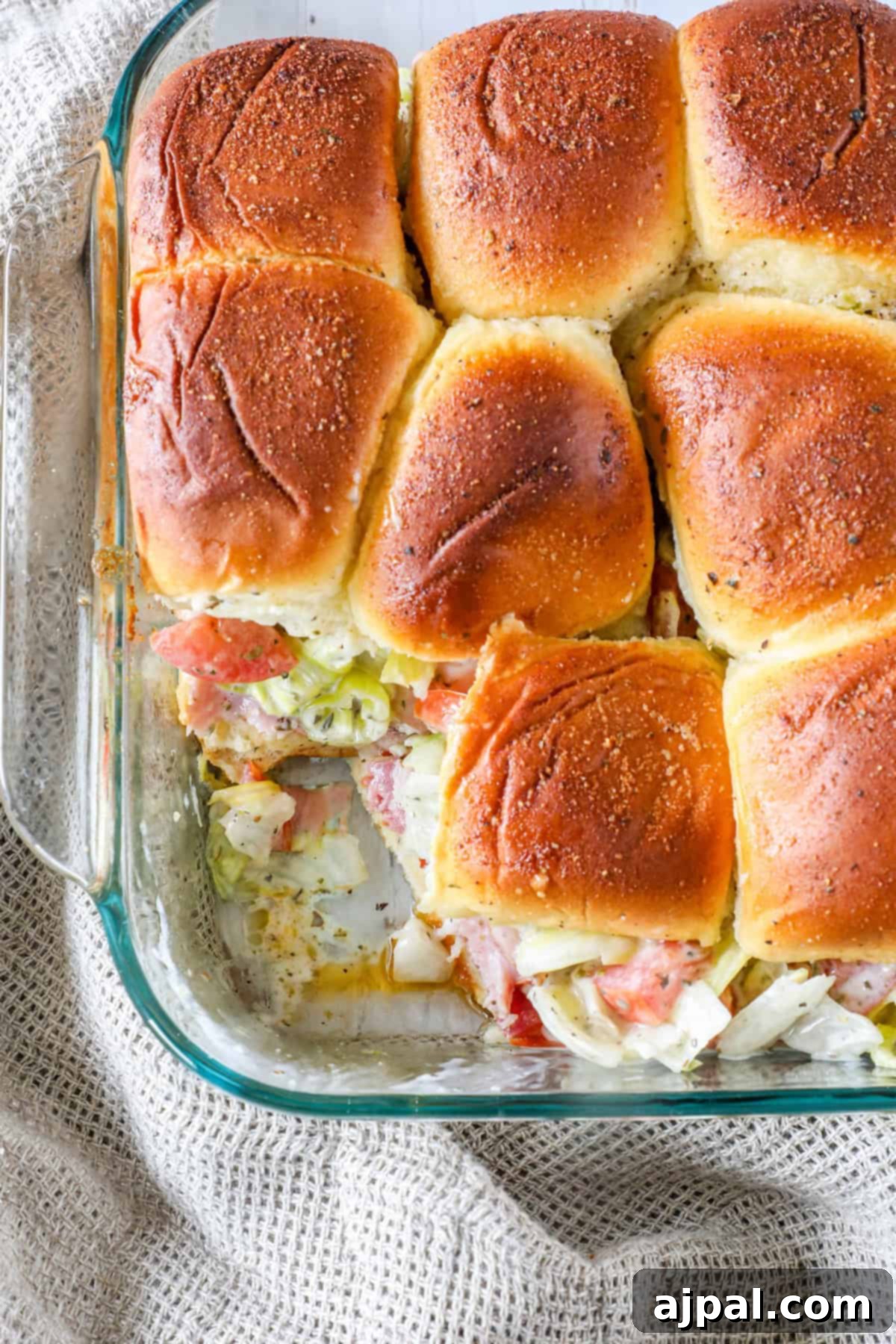 A glass baking dish contains baked sandwich sliders, with some missing, on a textured fabric surface.