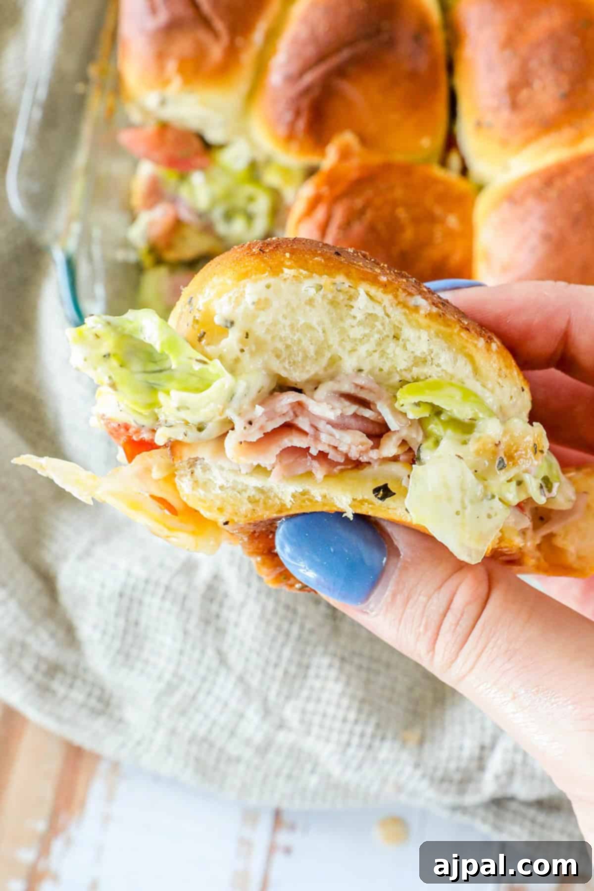 A hand holding a sandwich with ham, lettuce, and cheese in a soft roll. More sandwiches in the background on a beige cloth.