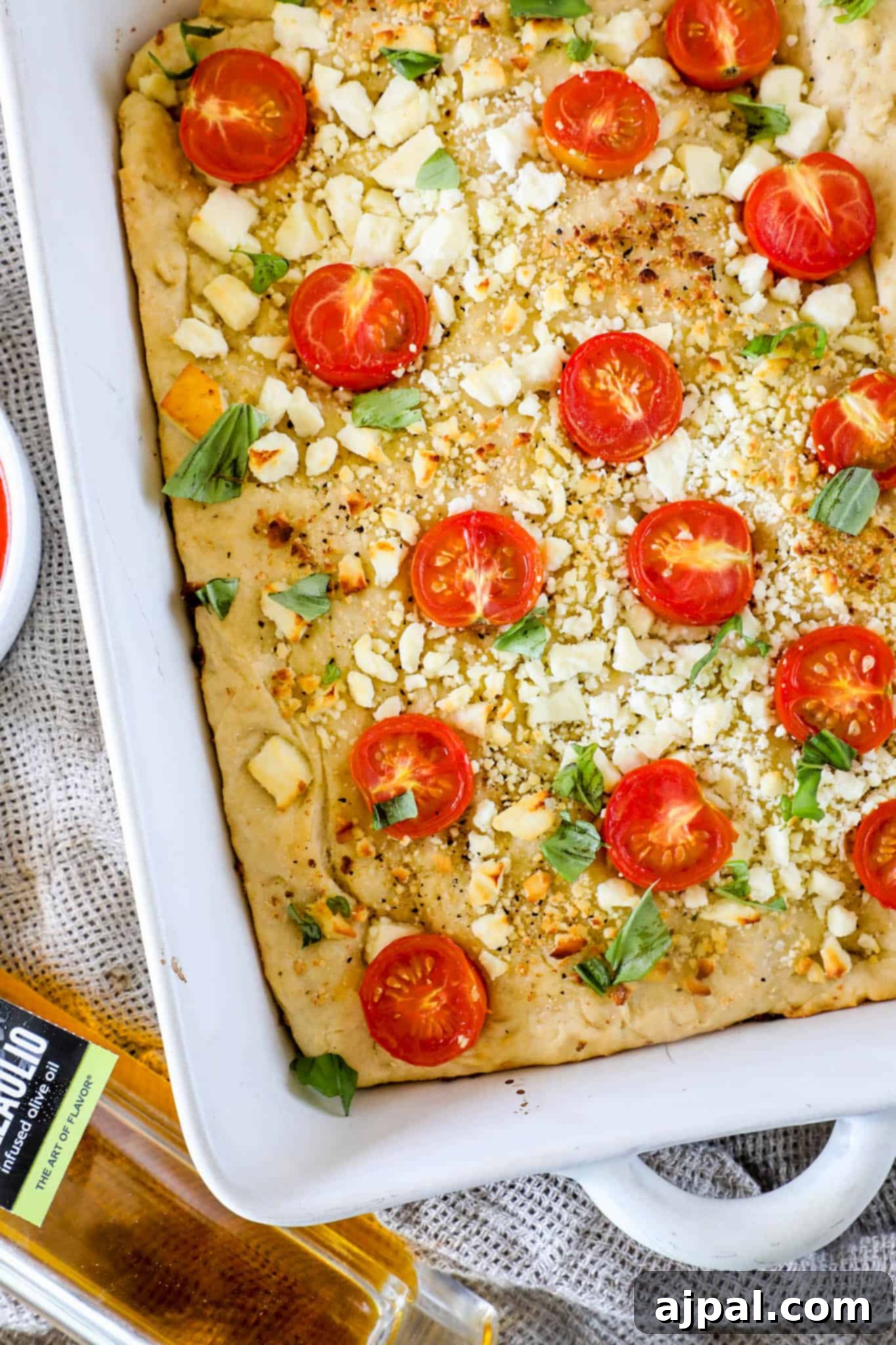 A rectangular pan of Mediterranean focaccia bread is adorned with vibrant cherry tomatoes, crumbled feta cheese, and fresh basil leaves, resting on a textured cloth backdrop.