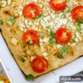Close-up of a baked Mediterranean focaccia bread topped with sliced tomatoes, crumbled cheese, and fresh basil leaves in a white baking dish.