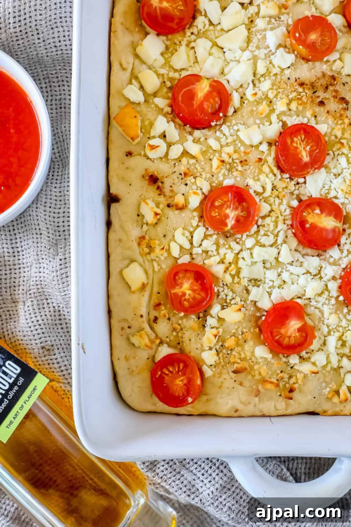 A rectangular pizza, reminiscent of Mediterranean focaccia bread, is topped with cherry tomato slices and crumbled cheese in a white dish. A small bowl of tomato sauce and a bottle of olive oil are nearby.