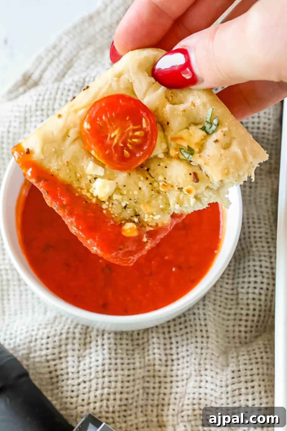 A hand holding a slice of Mediterranean focaccia bread with tomato and herbs is about to dip it into a bowl of red sauce on a textured surface.