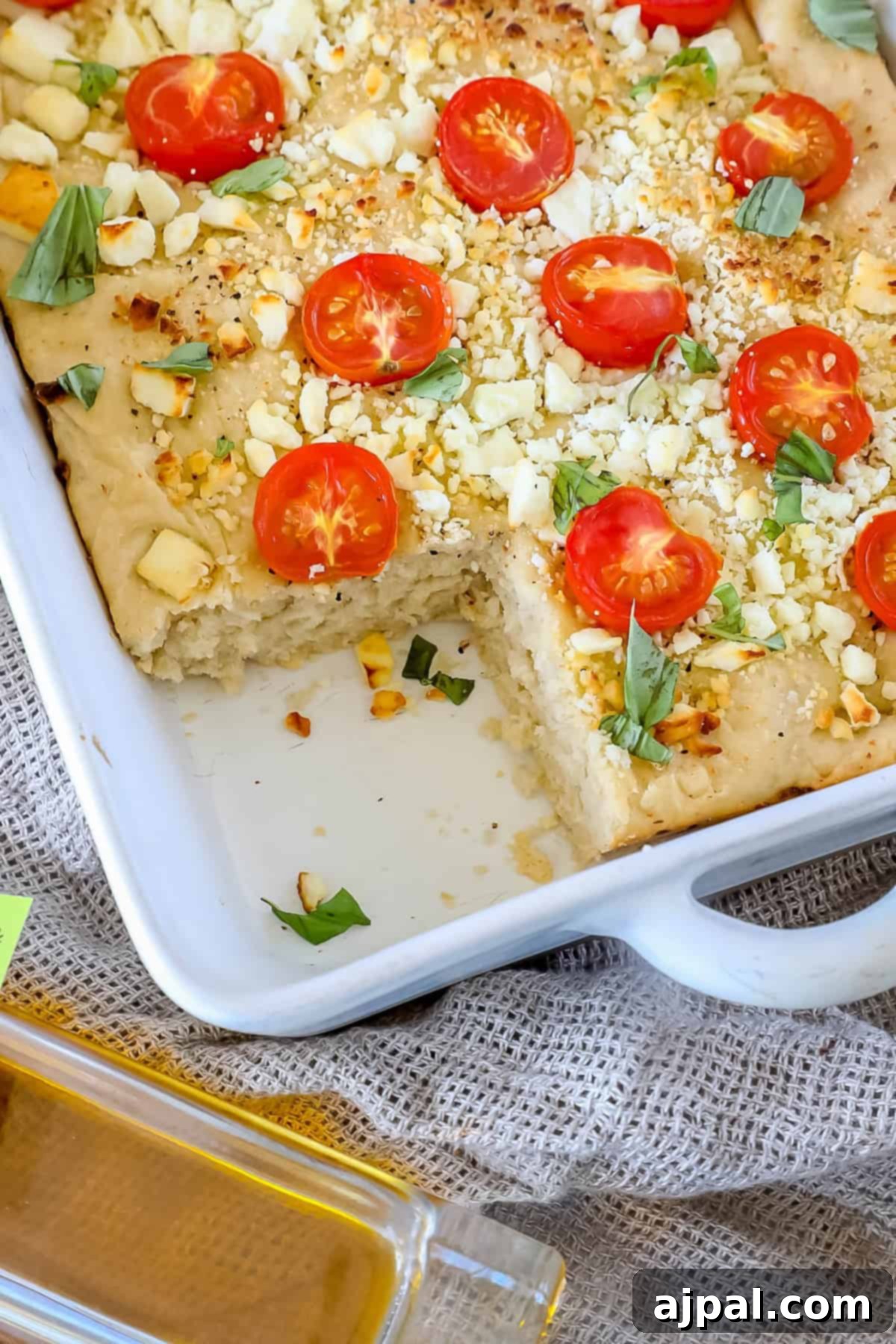 A close-up of Mediterranean focaccia bread topped with cherry tomatoes, cheese, and herbs in a white baking dish. A piece is missing, revealing the soft interior. A dish of olive oil is visible in the foreground.