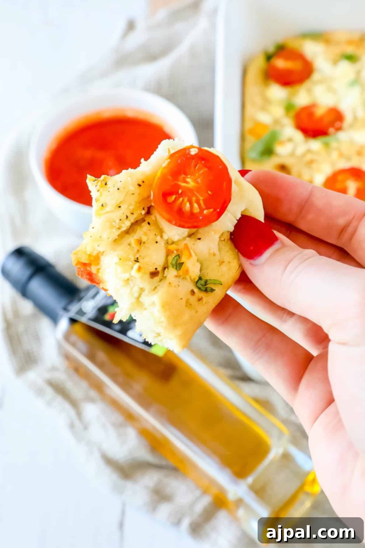 A hand holds a piece of Mediterranean focaccia bread, topped with juicy cherry tomatoes and fragrant herbs, while a bottle of olive oil and a bowl of rich red sauce linger invitingly in the background.