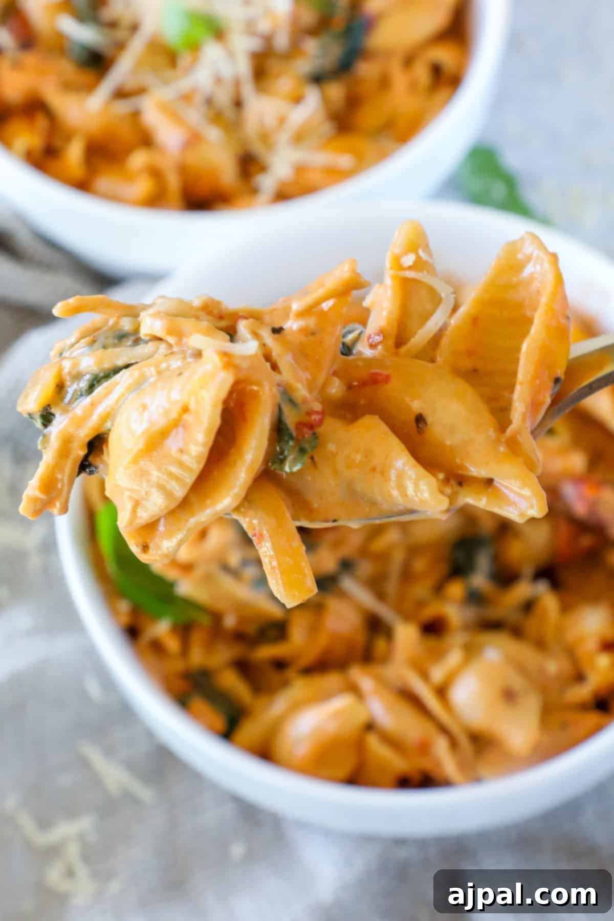 A close-up shot of a spoon lifting creamy pasta shells with spinach and sun-dried tomatoes, held above a generously filled bowl of the delicious dish.