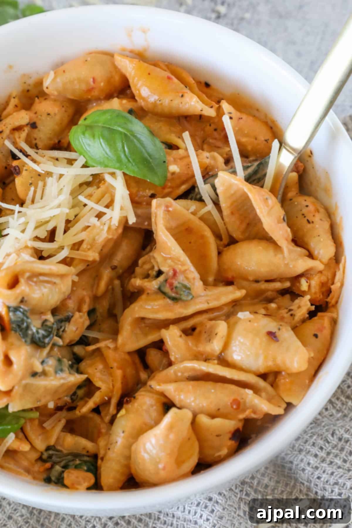 A heartwarming bowl of creamy pasta shells with fresh spinach, elegantly topped with grated cheese and a vibrant basil leaf, accompanied by a fork for immediate enjoyment.