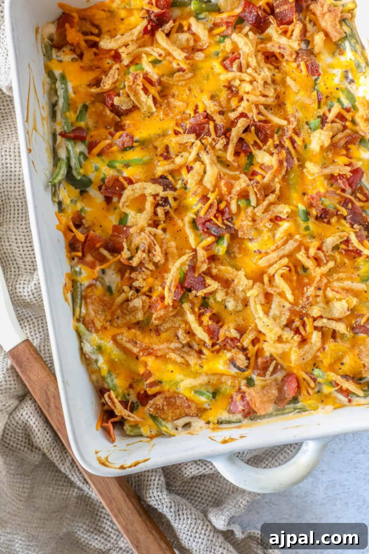 Ultimate Green Bean Bake 2 A green bean casserole baked to perfection, featuring melted cheese, crispy onions, and tender green beans, all nestled in a rectangular dish on a textured cloth.