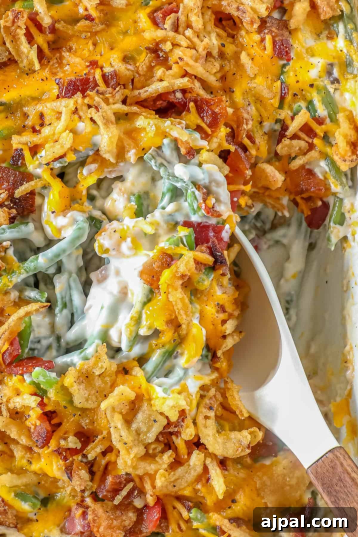Ultimate Green Bean Bake 3 A spoon serving a portion of green bean casserole reveals a close-up of the dish, topped with melted cheese, crispy fried onions, and savory bacon bits.