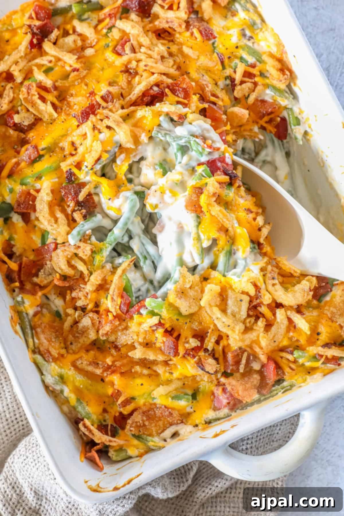 Ultimate Green Bean Bake 4 A casserole dish features a classic green bean casserole topped with melted cheese, crispy fried onions, and bacon, while a white serving spoon elegantly scoops a generous portion.