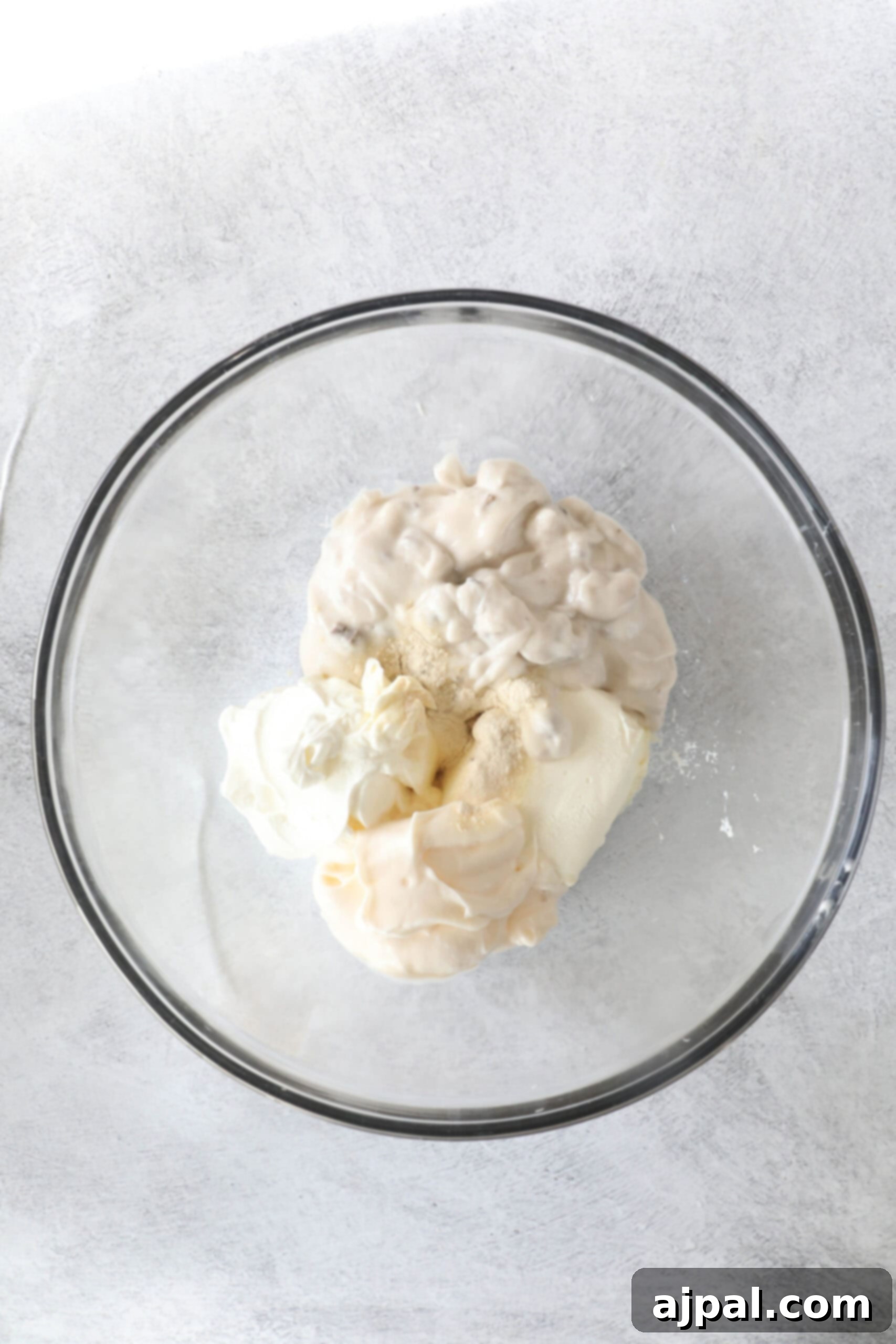 Ultimate Green Bean Bake 5 A glass bowl containing dollops of mayonnaise, sour cream, and a sprinkle of garlic powder sits on a gray surface, ready to enhance the flavors of your green bean casserole.