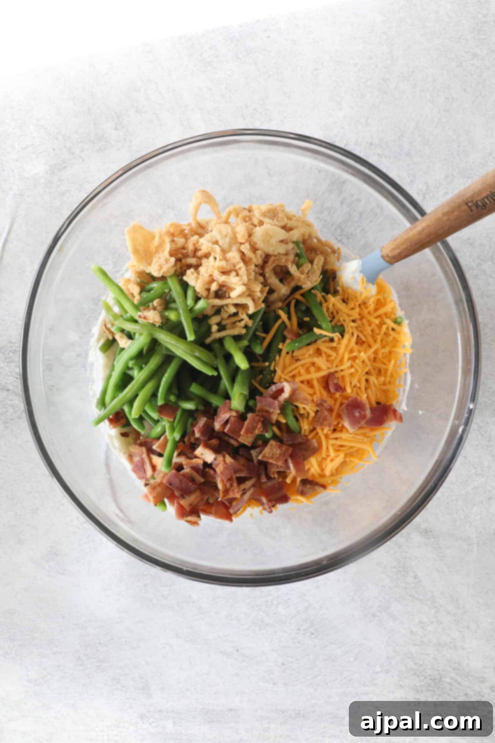 Ultimate Green Bean Bake 6 In a glass bowl, this green bean casserole features vibrant green beans, shredded cheese, crispy onions, chopped bacon, and a spoon for easy serving.