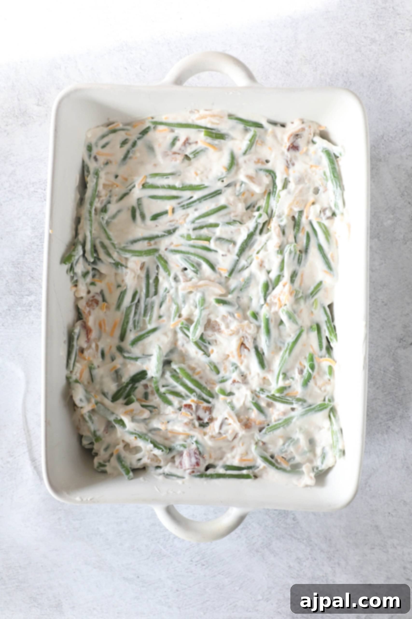 Ultimate Green Bean Bake 7 A classic green bean casserole dish filled with a creamy mixture and topped with shredded cheese sits elegantly on a light background.