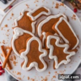 A plate of gingerbread cookies, artfully shaped into trees and bells, decorated with intricate white icing. Two cinnamon sticks rest beside the gingerbread cookies, adding a touch of aromatic spice.