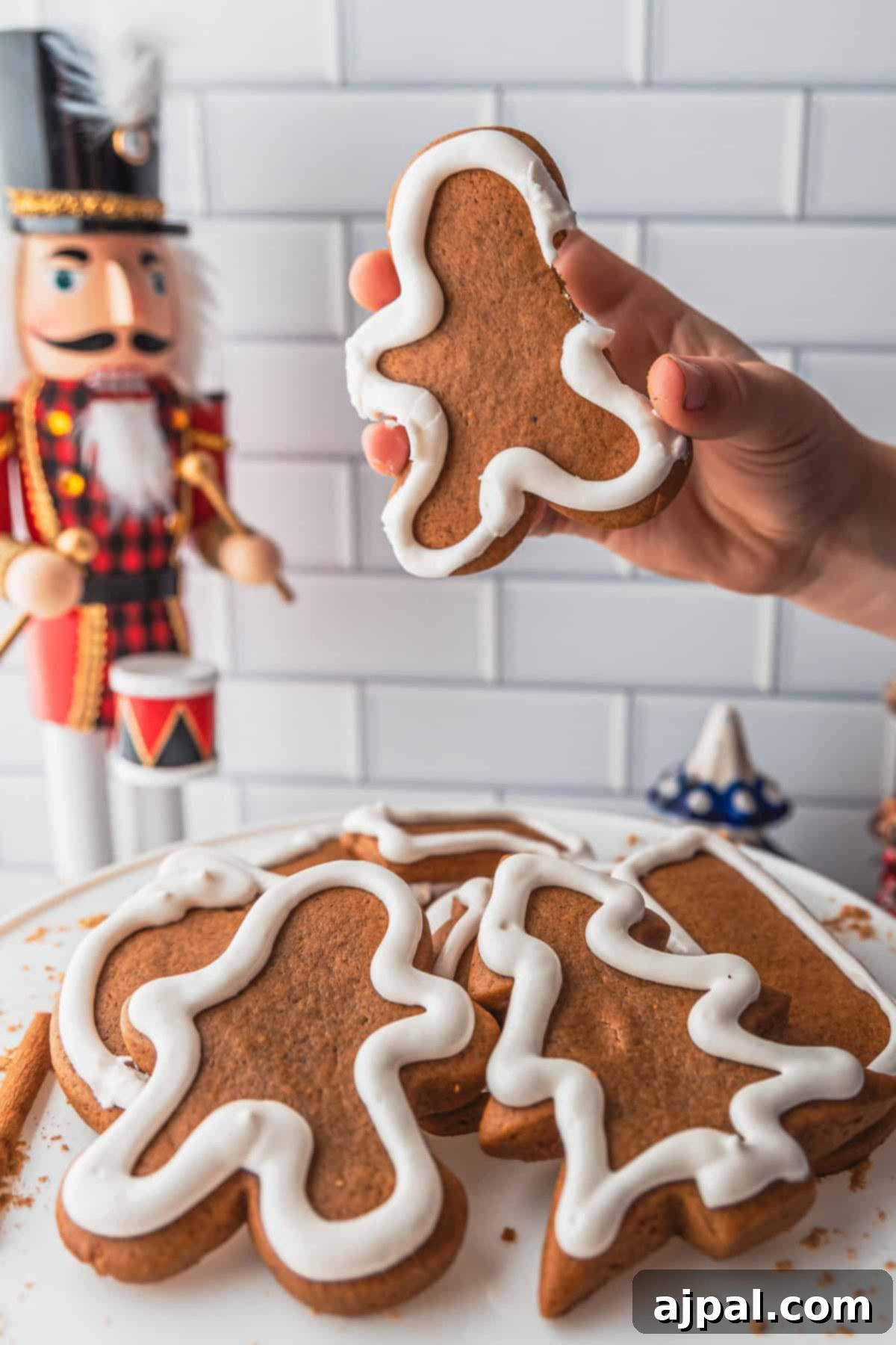 Perfectly Spiced Gingerbread 4 A hand holds a gingerbread cookie with white icing, hovering above a plate of gingerbread cookies, while a nutcracker figure stands watchful in the background.