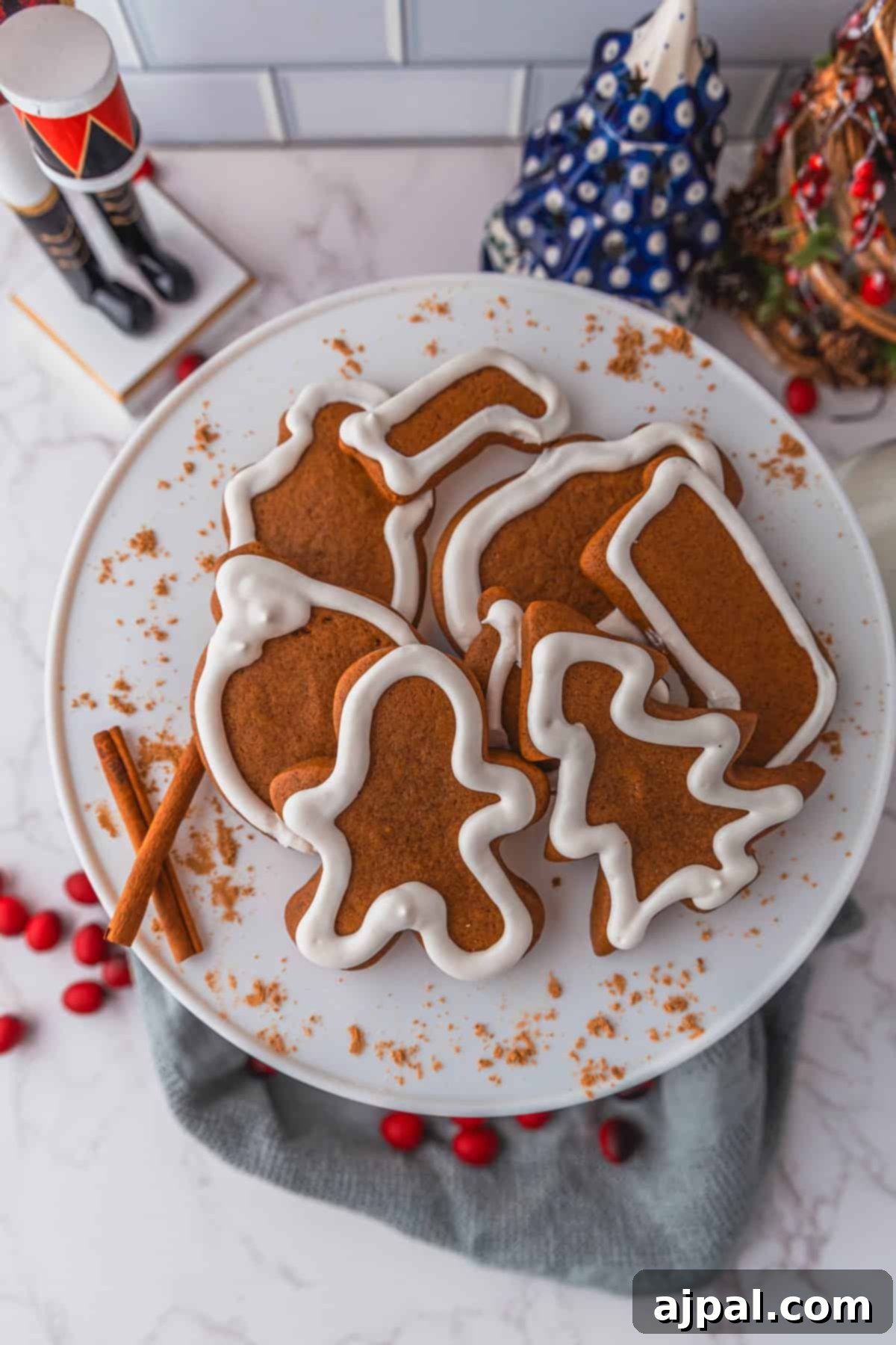 Perfectly Spiced Gingerbread 6 A plate of gingerbread cookies with white icing, shaped like a mitten, stocking, gingerbread person, bell, and tree sits invitingly on a table adorned with cinnamon sticks and red berries.