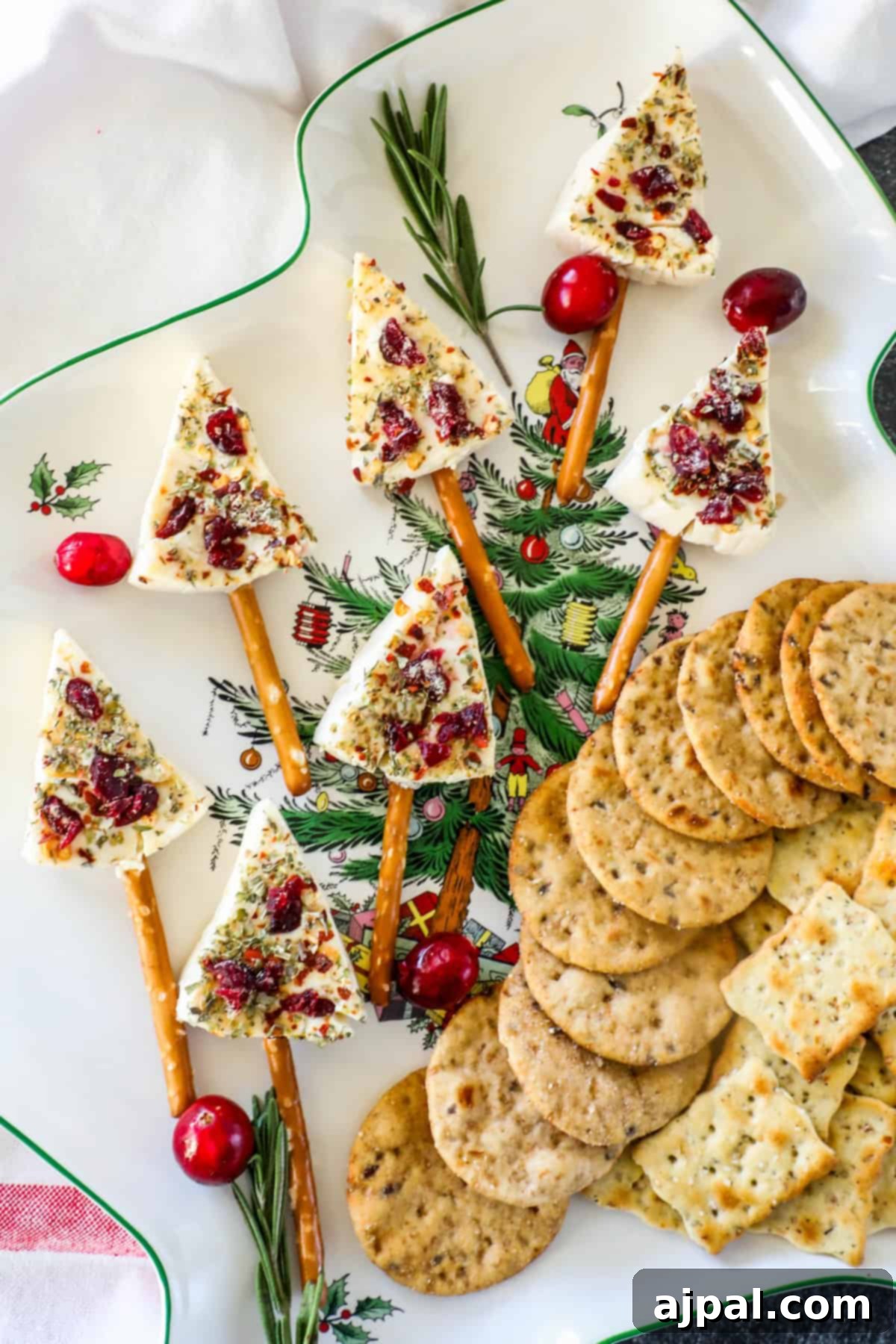 Festive Cheese Tree Bites 2 Festive appetizer plate showcasing delightful Christmas tree cheese wedges, expertly placed on pretzel sticks, adorned with vibrant fresh herbs and tart cranberries. The presentation is completed with an assortment of gourmet crackers and aromatic rosemary sprigs, perfect for holiday gatherings.