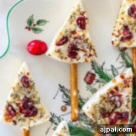 Festive Cheese Tree Bites 7 Festive tree-shaped treats and Christmas tree cheese wedges with cranberries and herbs embellish a decorative plate, featuring pretzel sticks as stems and a small red ornament for decoration.