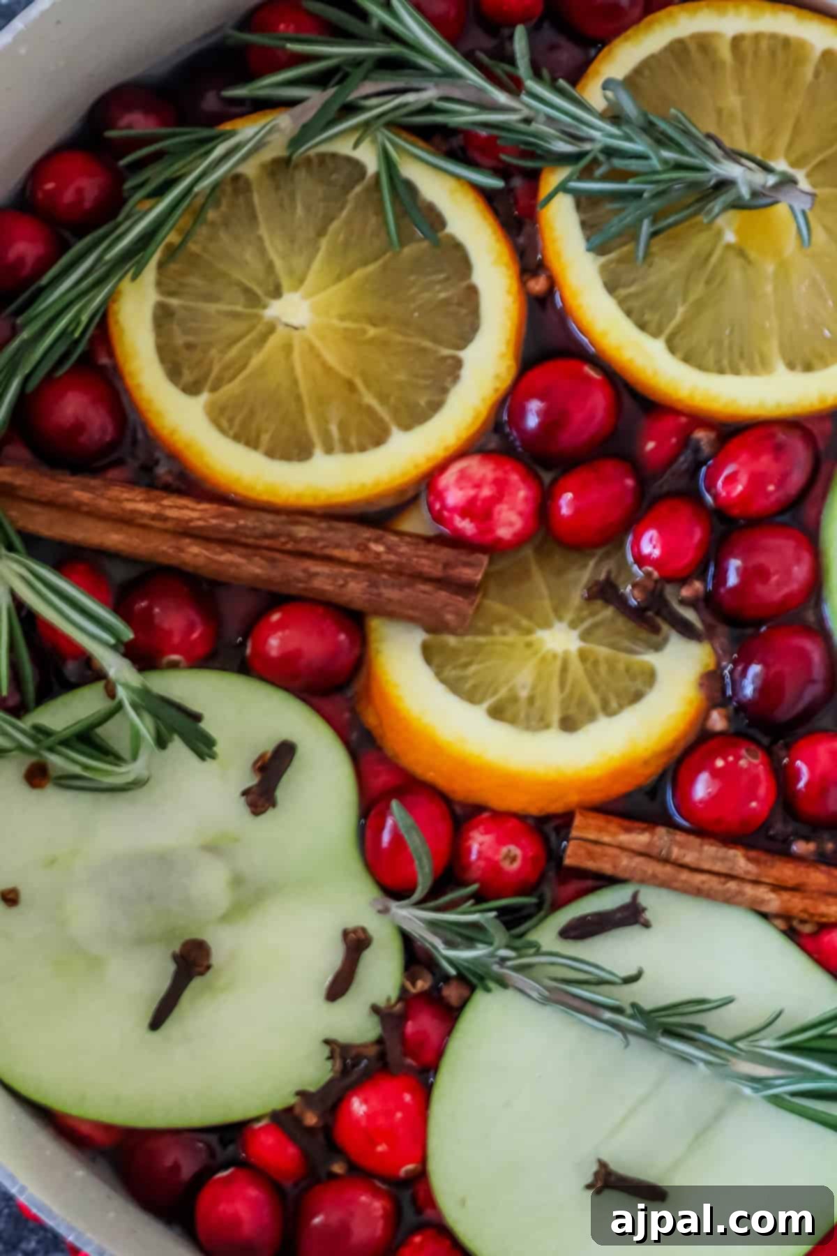 A pot filled with sliced oranges, apples, cranberries, cinnamon sticks, sprigs of rosemary, and cloves in liquid creates a festive Christmas potpourri, radiating warmth and holiday spirit.