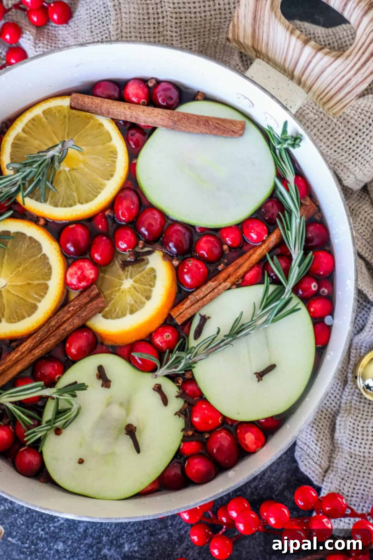 A festive Christmas potpourri overflows from a pot filled with cranberries, sliced oranges and apples, cinnamon sticks, rosemary sprigs, and cloves. It sits on a table draped with a beige cloth, surrounded by scattered red berries, creating a warm and inviting holiday display.